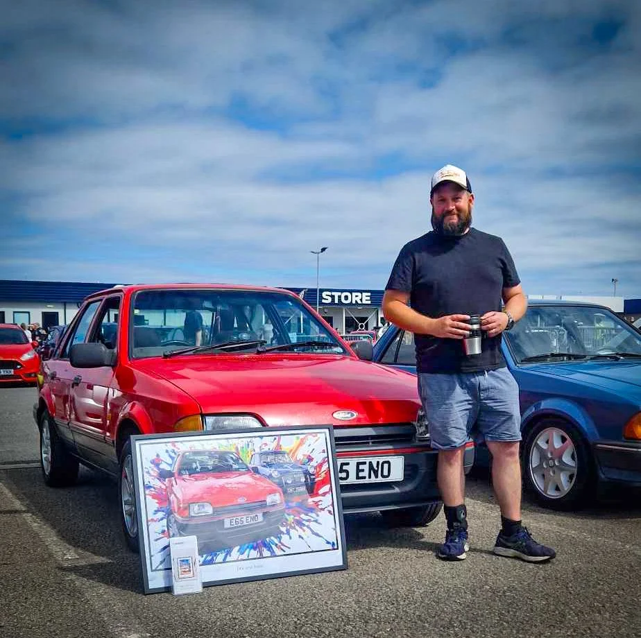 Man standing in parking lot next to a red vintage car, holding a travel mug, with a framed colorful artwork of the car and a store building in the background.