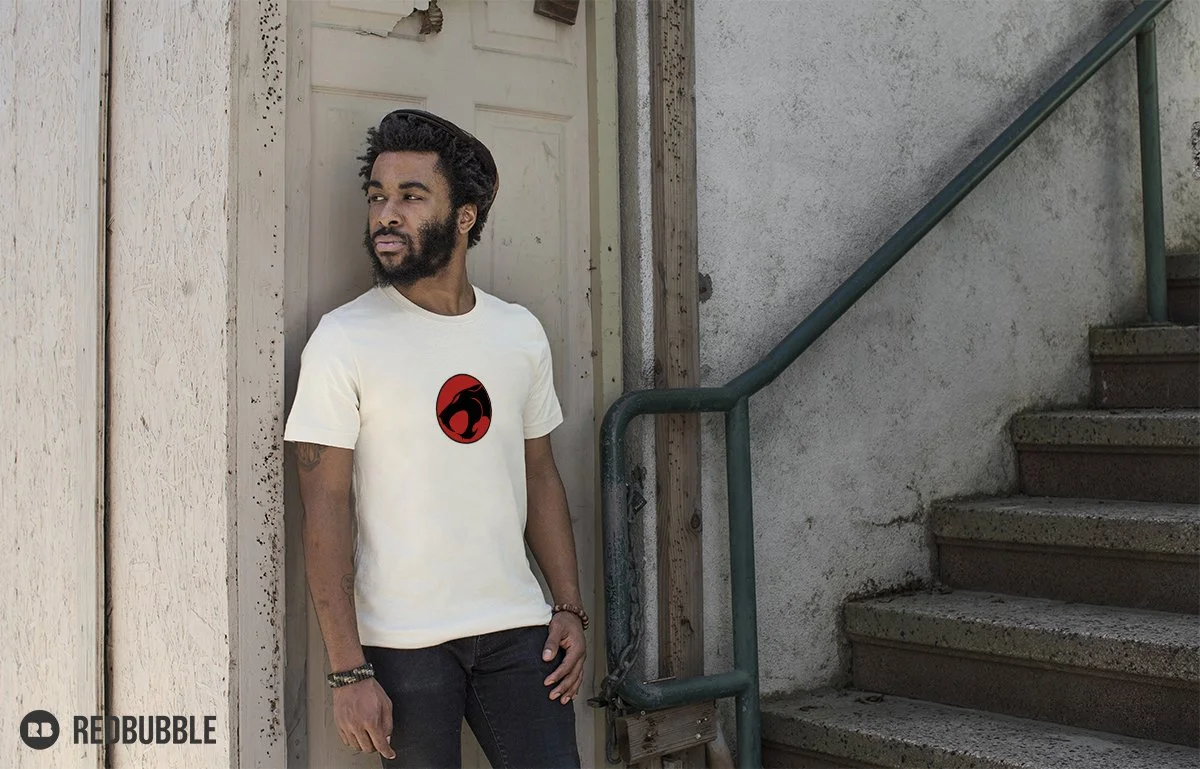 A man with a beard and black hair standing next to a stairway outside, wearing a white T-shirt with a red and black logo on it.