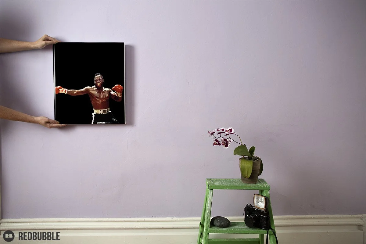Two hands are holding a framed photograph of a boxer with red gloves, a muscular build, and a clenched fist, with a black background and a few signatures at the bottom.