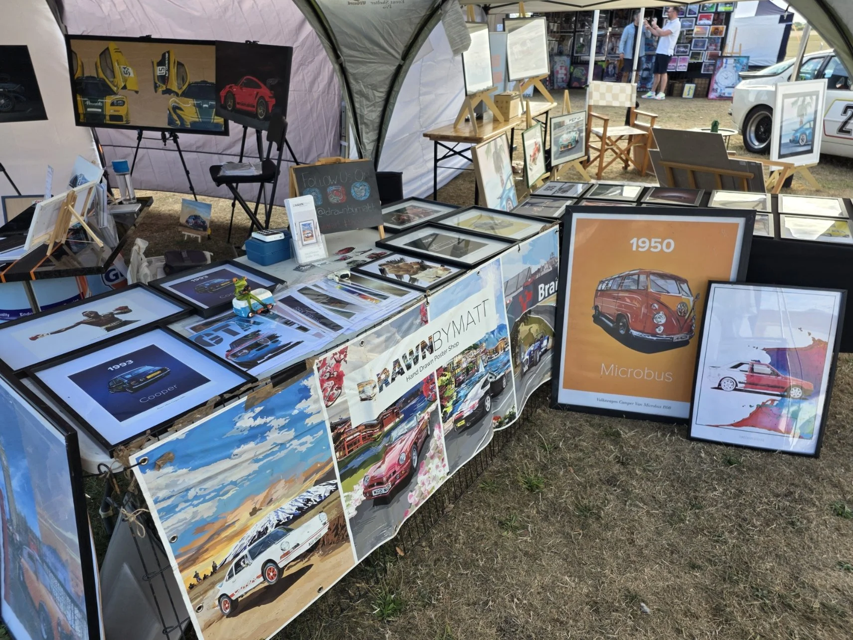 At an outdoor art and photography stall, framed pictures of cars and vintage vehicles are displayed on tables and walls, with some artwork featuring a red classic car, a white vintage car, and a blue sports car. There is a large framed poster of a 19