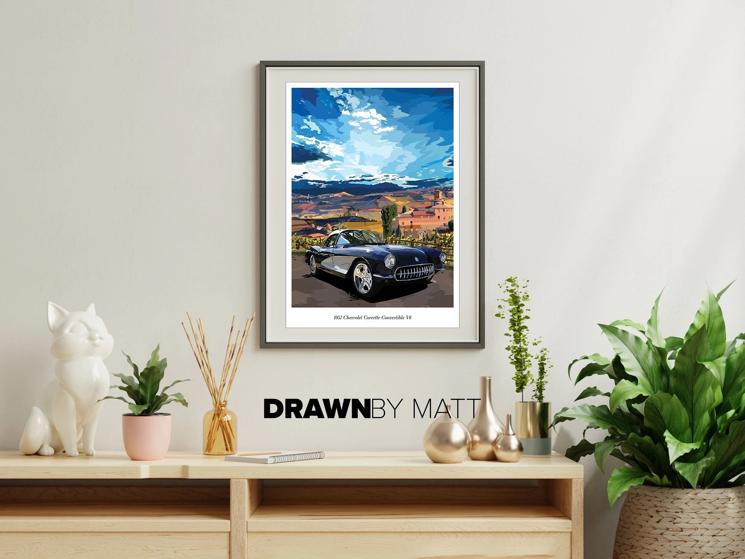 Framed digital art print of a 1957 Chevrolet Corvette Convertible V8 with a scenic sky and landscape in the background, hanging on a white wall above a wooden table decorated with plants, vases, and a cat figurine.