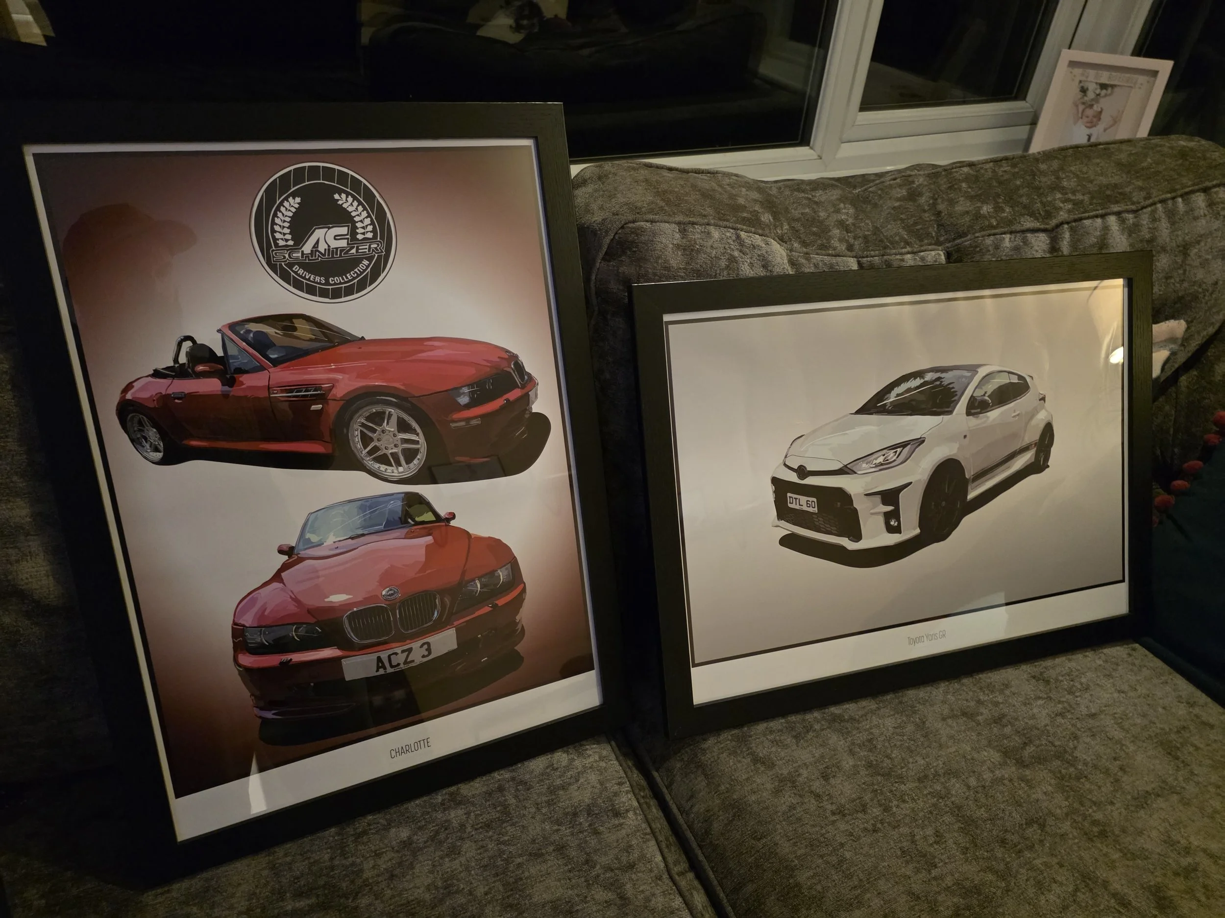 Two framed car posters leaning against a sectional sofa on a carpeted floor, with a window and family photos in the background.
