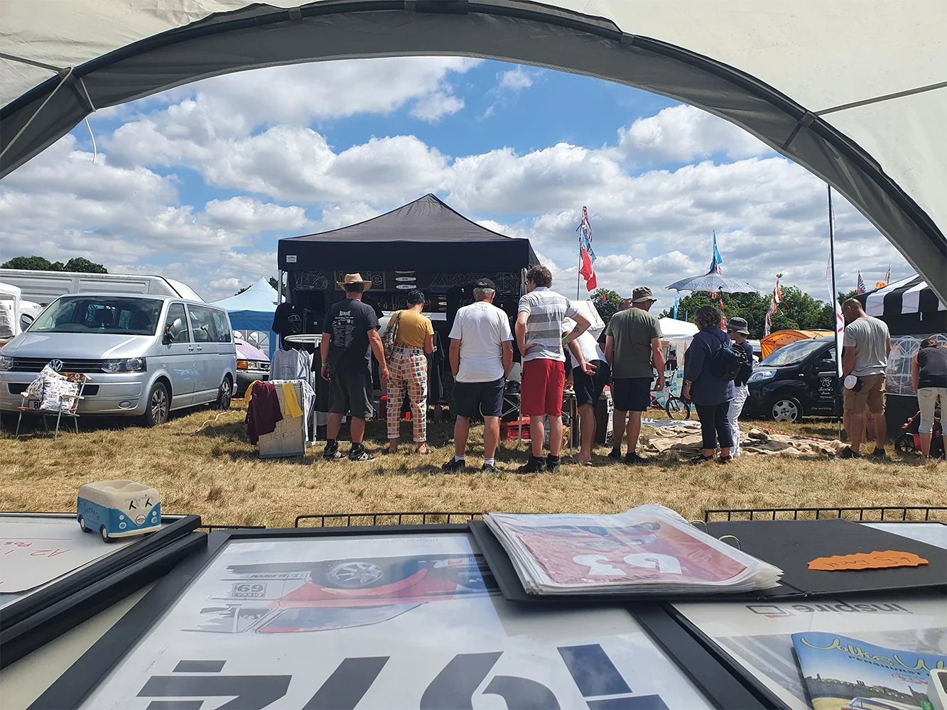 People gathered at an outdoor event under tents, with vehicles and flags visible, seen from inside a tent or booth with brochures and a small toy on the table.