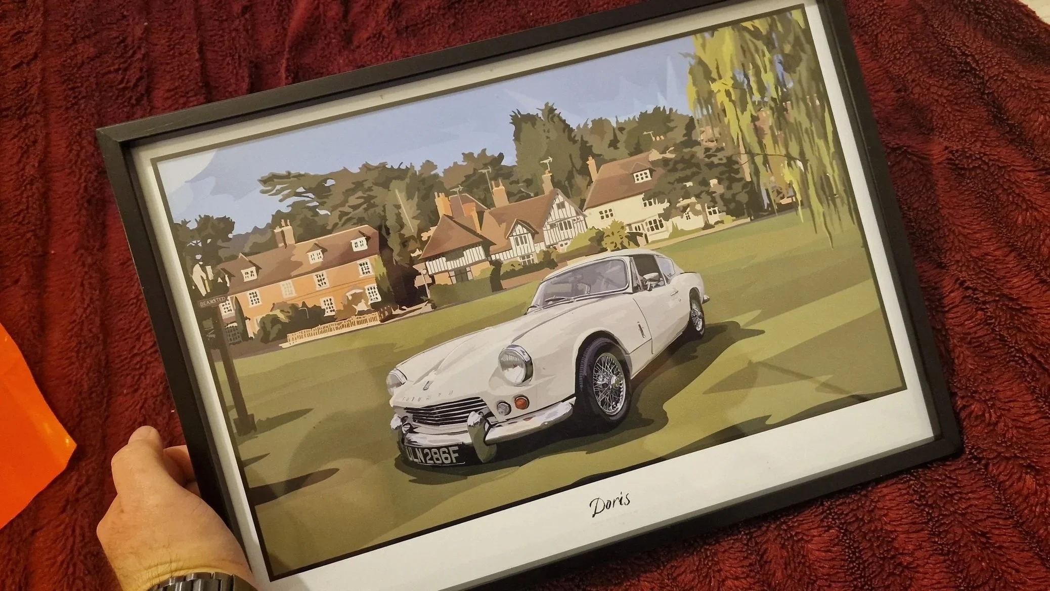 Digital illustration of a classic white car, parked on a lawn, with a background of a village with houses and trees, framed with the name 'Doris' at the bottom.