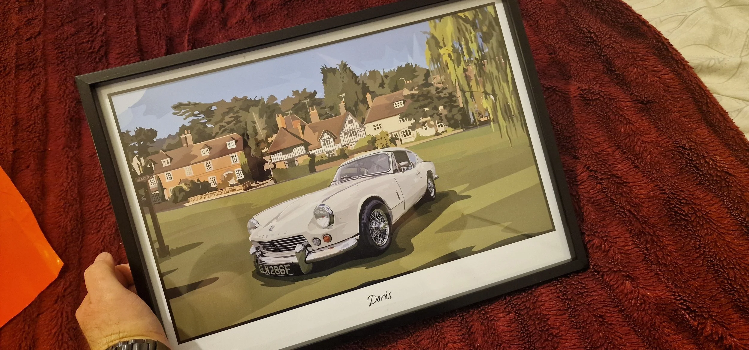 Framed digital illustration of a vintage white sports car parked on a lawn in front of a large house with trees in the background, held by a hand, on a red textured surface.