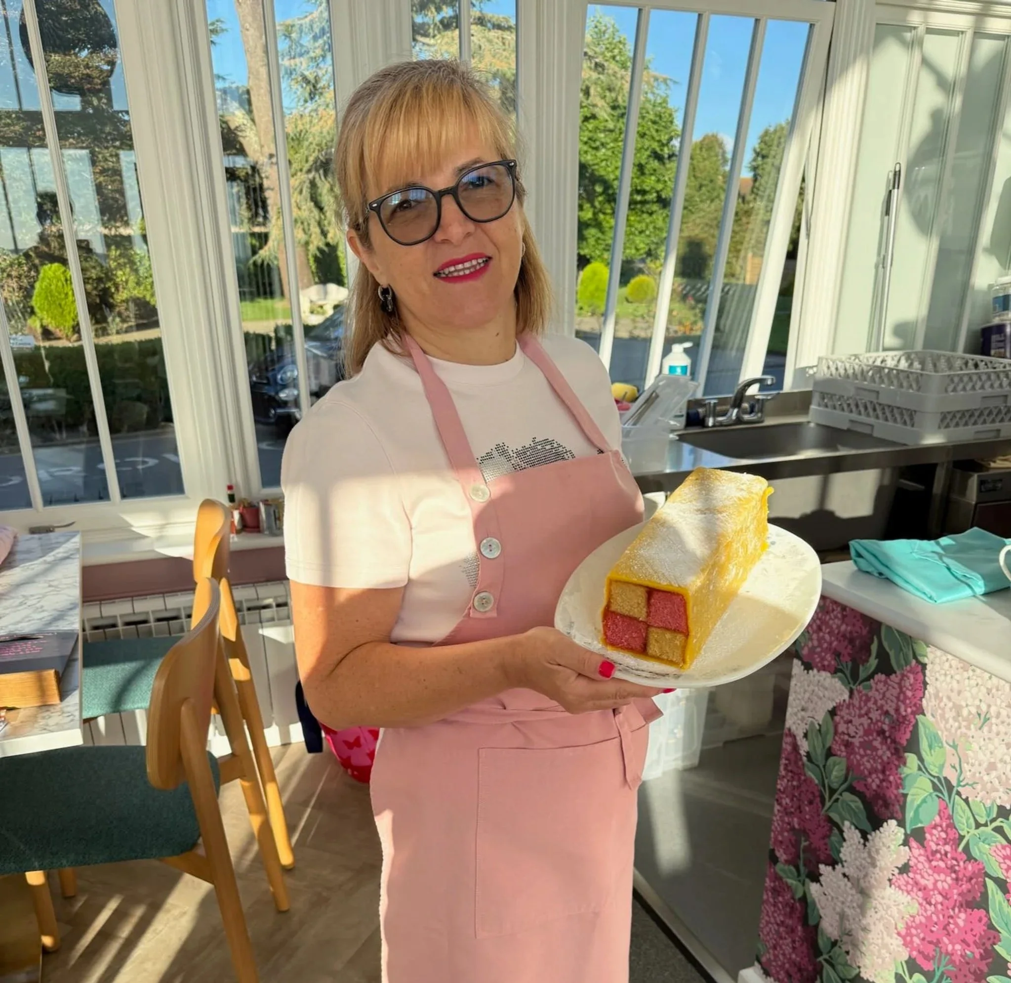 A woman with blonde hair, glasses, and red lipstick wearing a white t-shirt and pink apron holding a plate with a large rectangular slice of yellow cake with pink filling, dusted with powdered sugar, in a sunlit kitchen with large windows, green trees outside, and floral tablecloth.