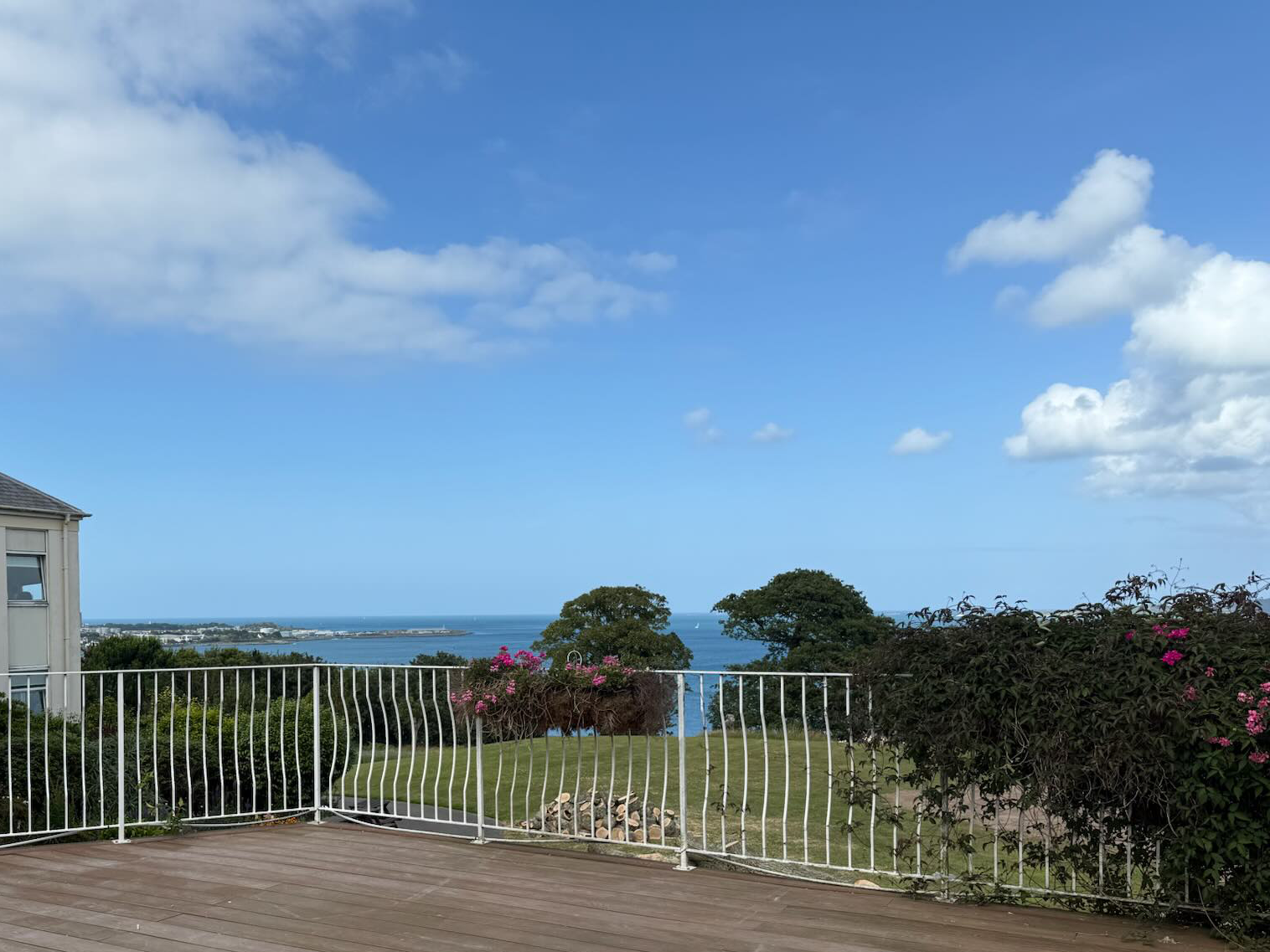 View of a coastal landscape with blue sky, clouds, trees, and a distant ocean seen beyond a white railing on a wooden deck.