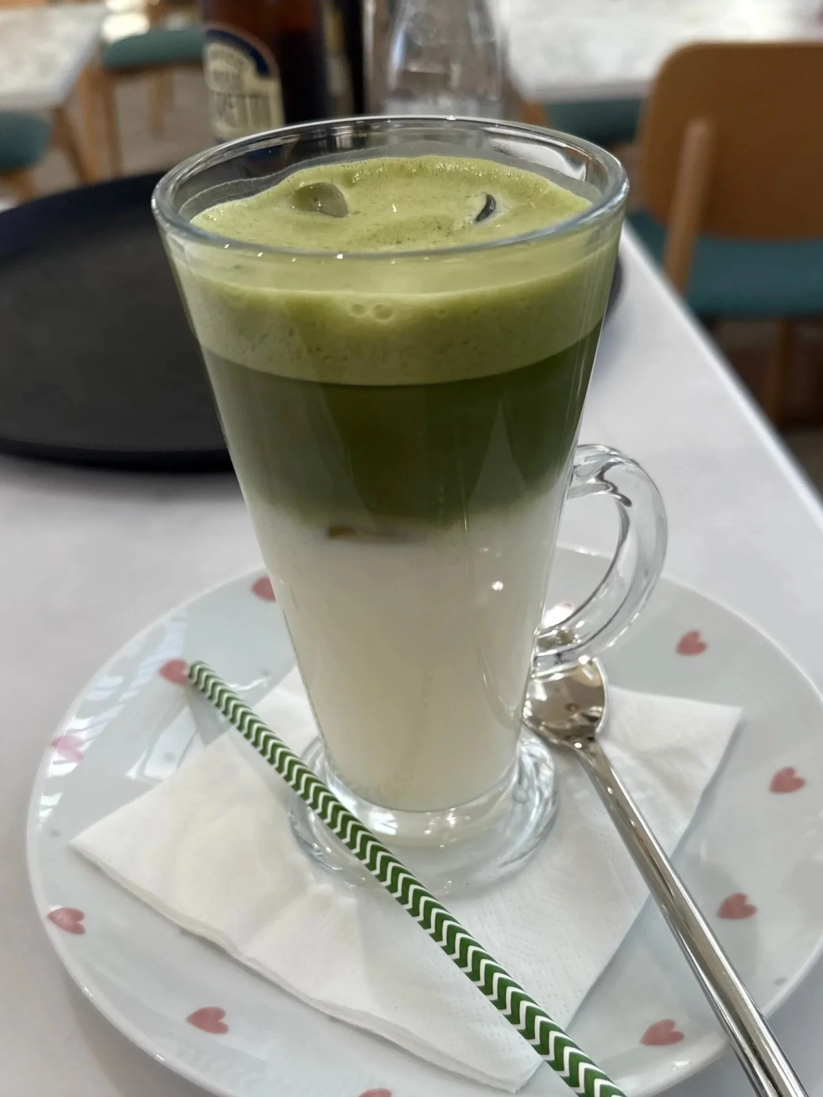 A tall glass with a layered green matcha drink topped with foam, served on a heart-patterned plate with a spoon, green and white straw, and a napkin.