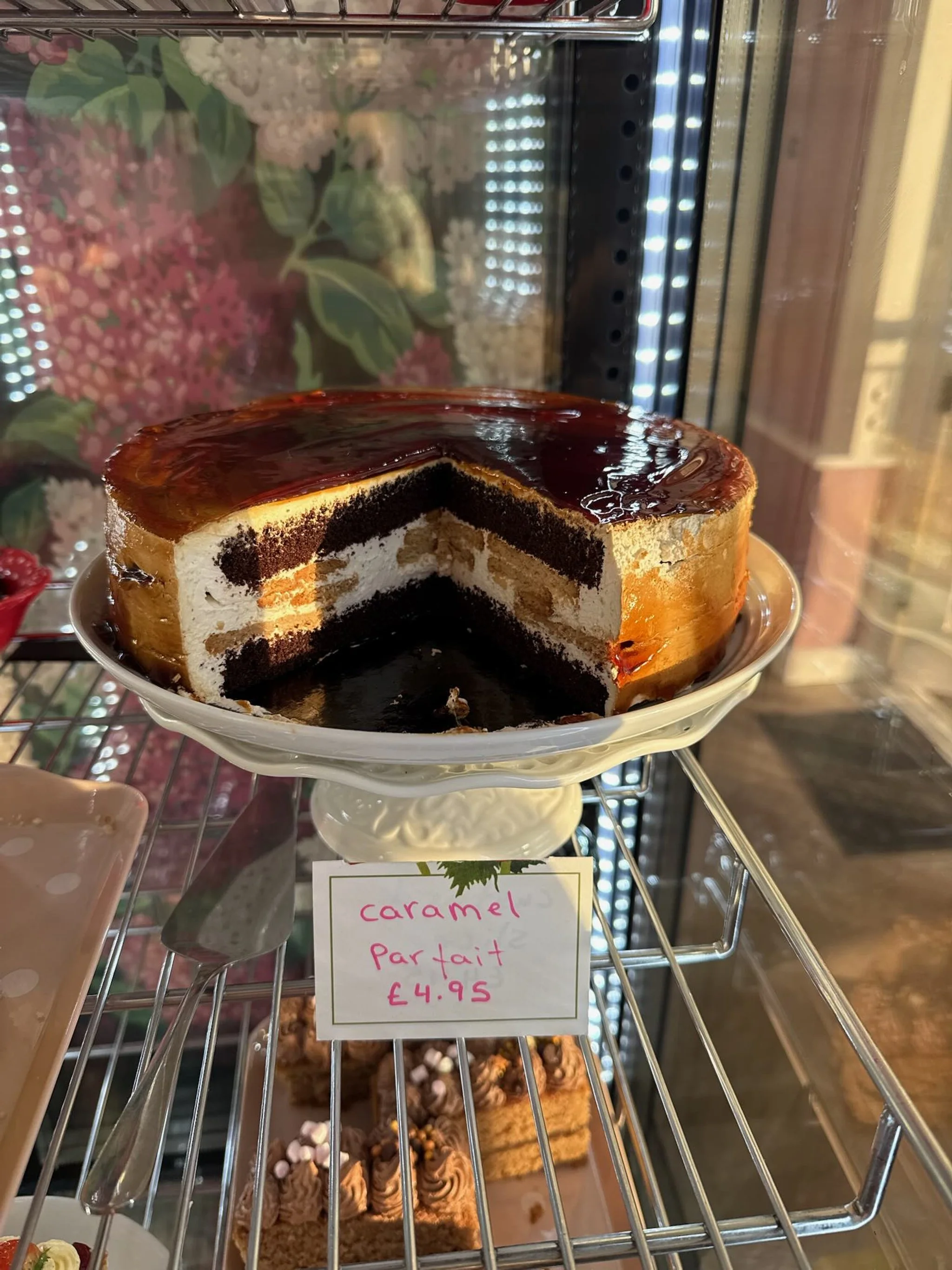 A caramel mille-feuille parfait cake with layers of cream and caramel on a white cake stand, inside a display case.