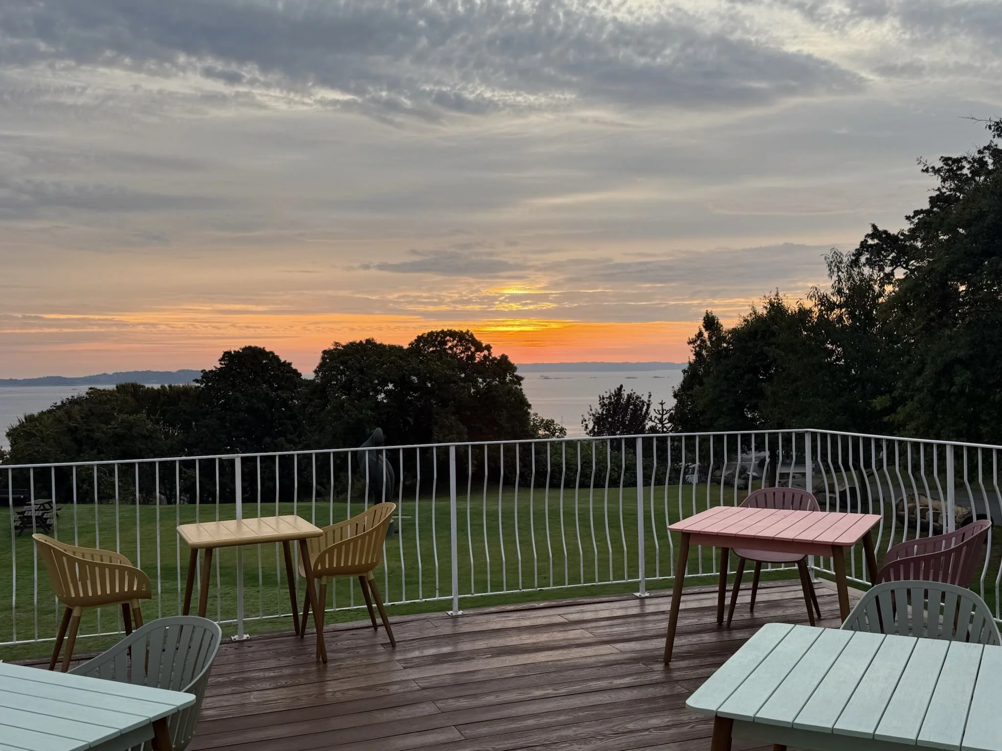 Sunset view over water with trees, seen from a wooden deck with outdoor tables and chairs and a metal railing.