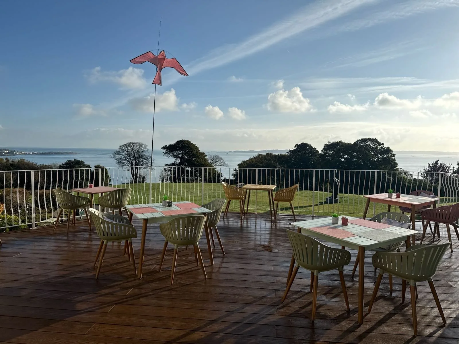 Outdoor dining area with wooden tables and chairs on a deck, overlooking a grassy area with trees and water in the distance, under a partly cloudy sky.