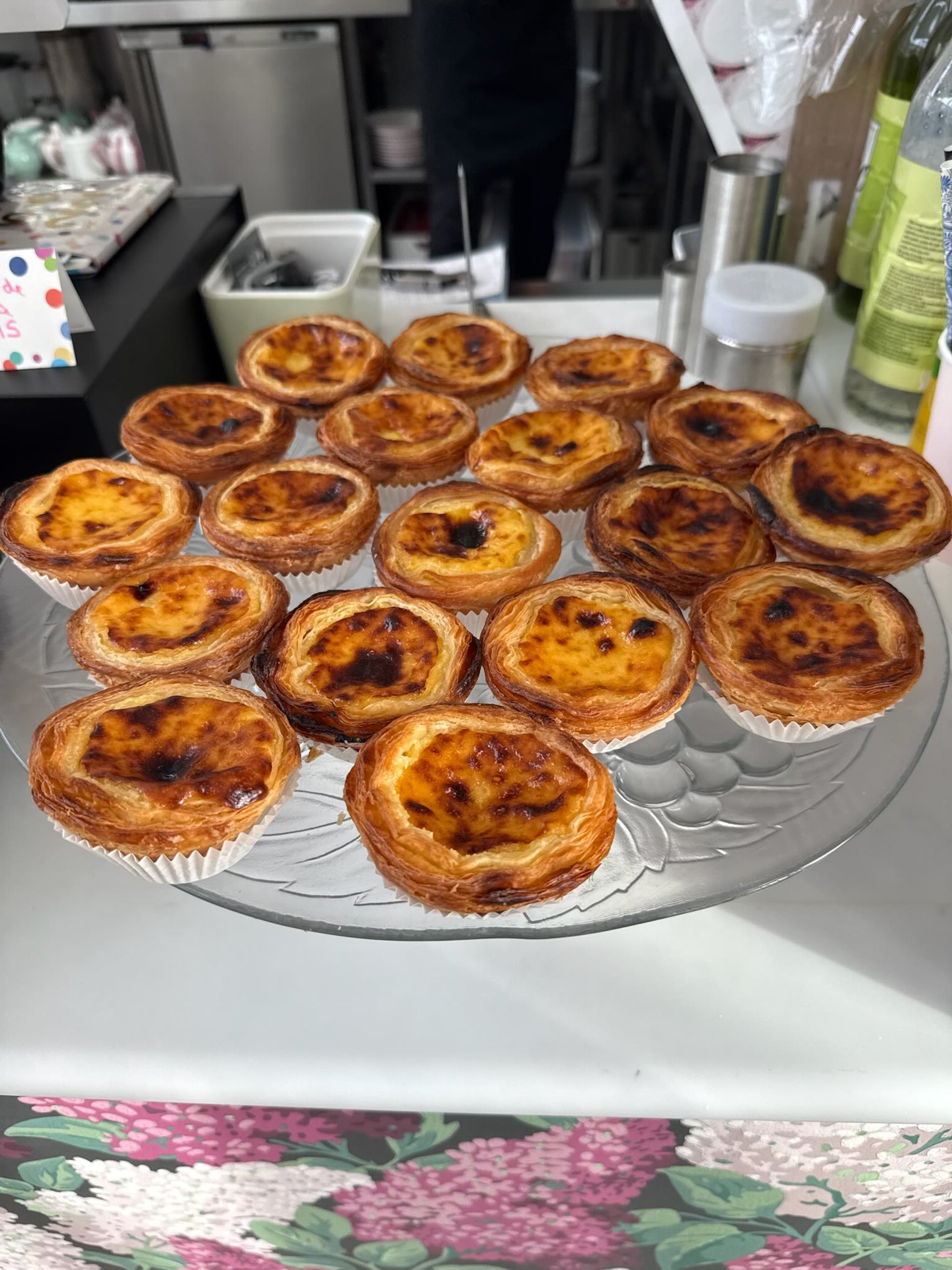 Tray of 20 freshly baked Portuguese custard tarts on a glass platter, with a busy kitchen in the background.
