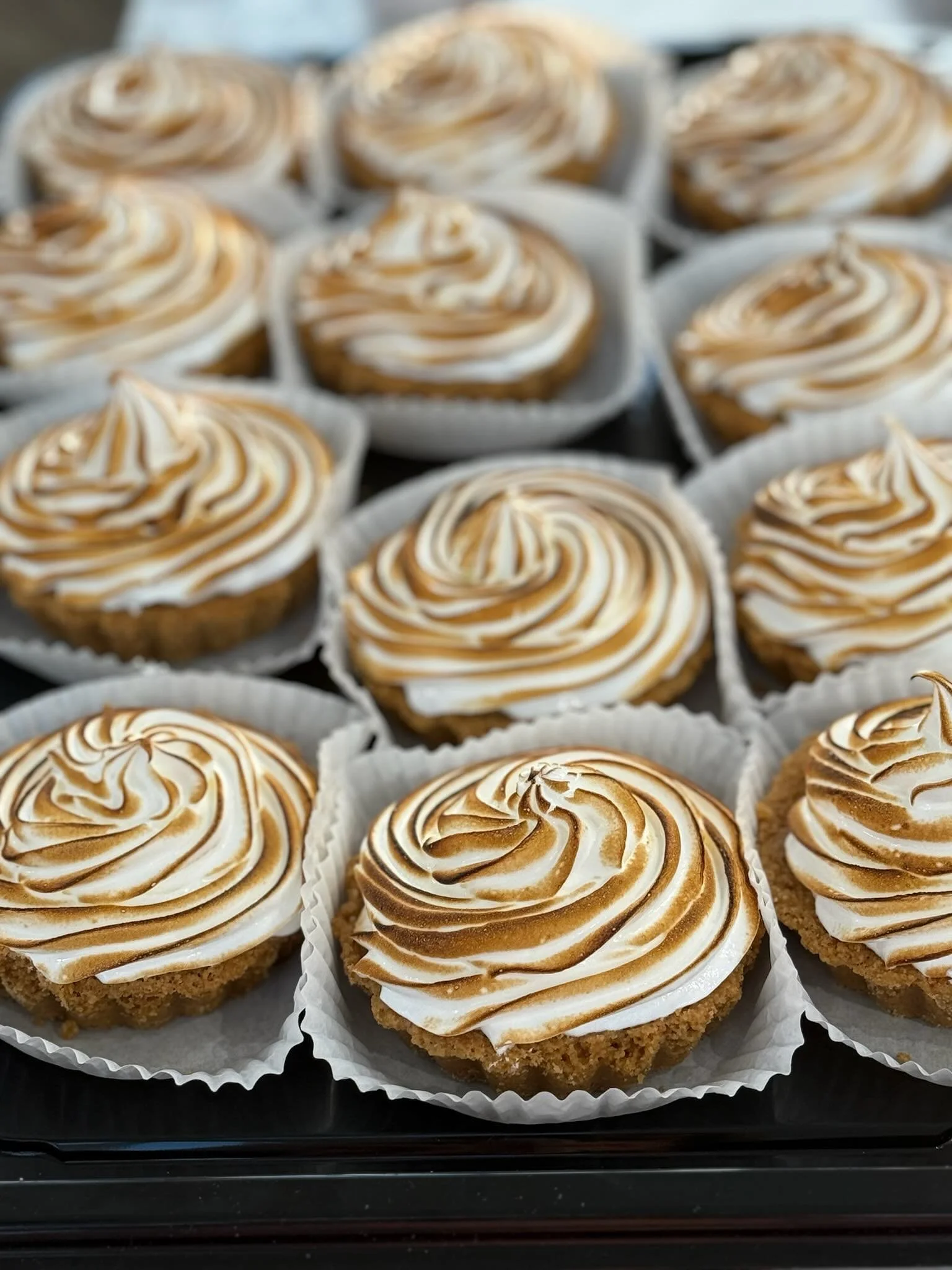 Close-up of vanilla cupcakes topped with swirls of toasted meringue in white paper cups on a black tray.