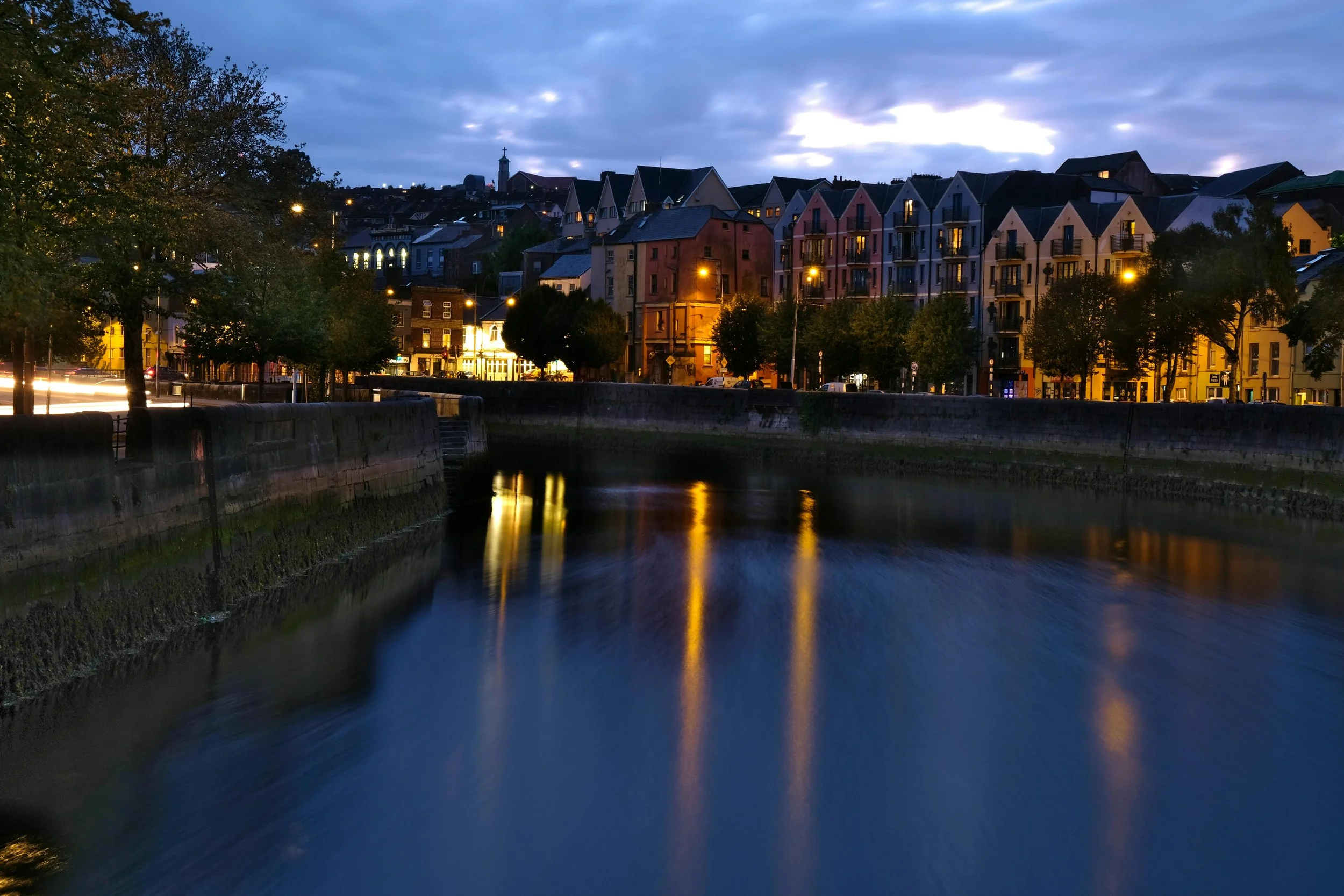 Nighttime view of a cityscape with colorful buildings along a river, illuminated street lights, and trees, with reflections on the water surface.