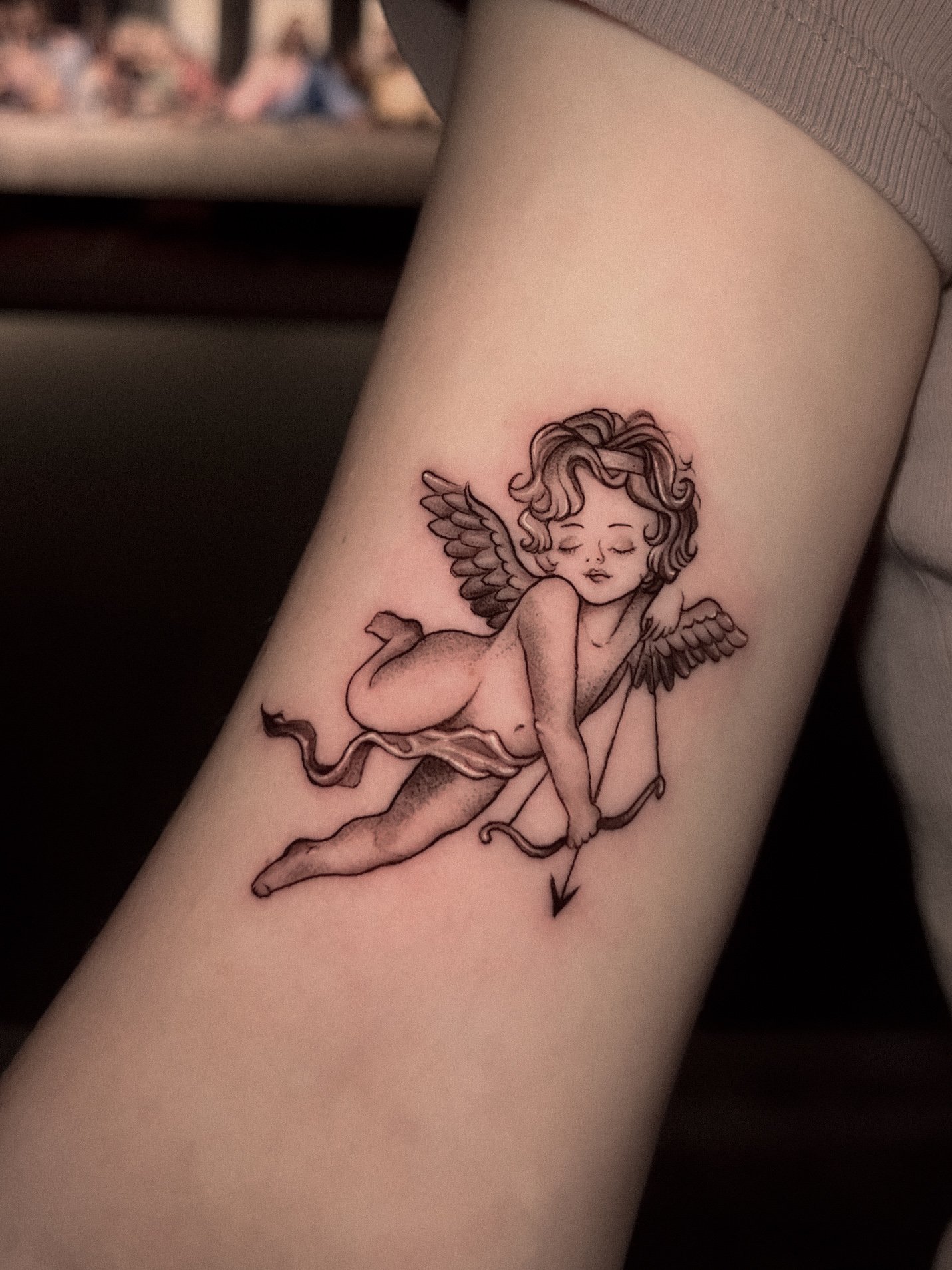 A tattoo of a cherub with wings, holding a bow and arrow, with eyes closed and a serene expression.