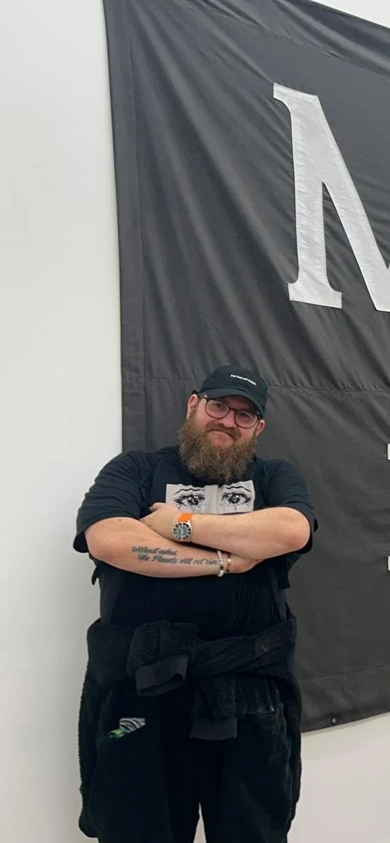Leslie Poet, wearing a black cap and T-shirt, standing with arms crossed in front of a large black banner with white lettering.