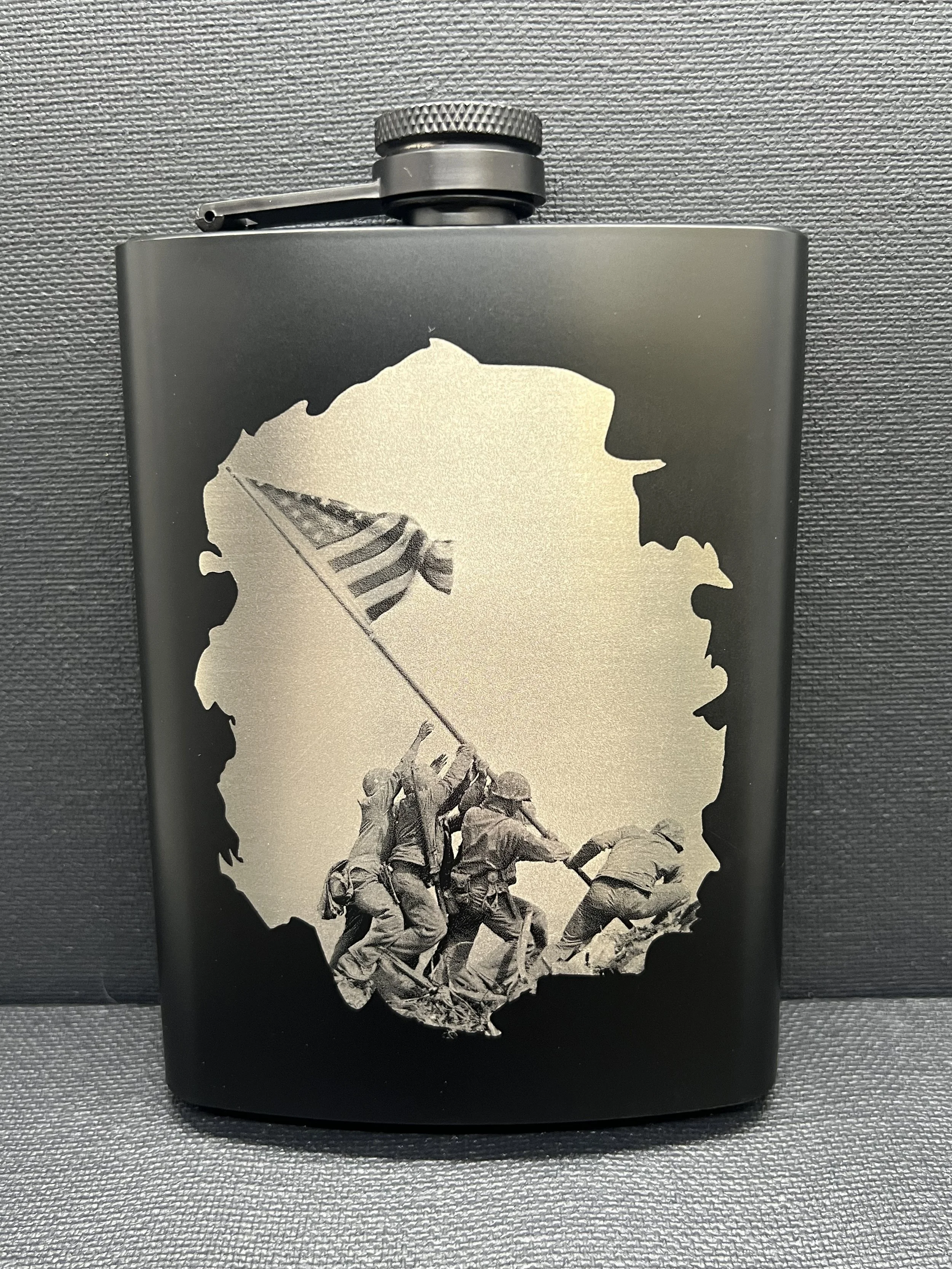 Black stainless steel hip flask engraved with the iconic Iwo Jima flag raising scene, shown against a textured dark background.