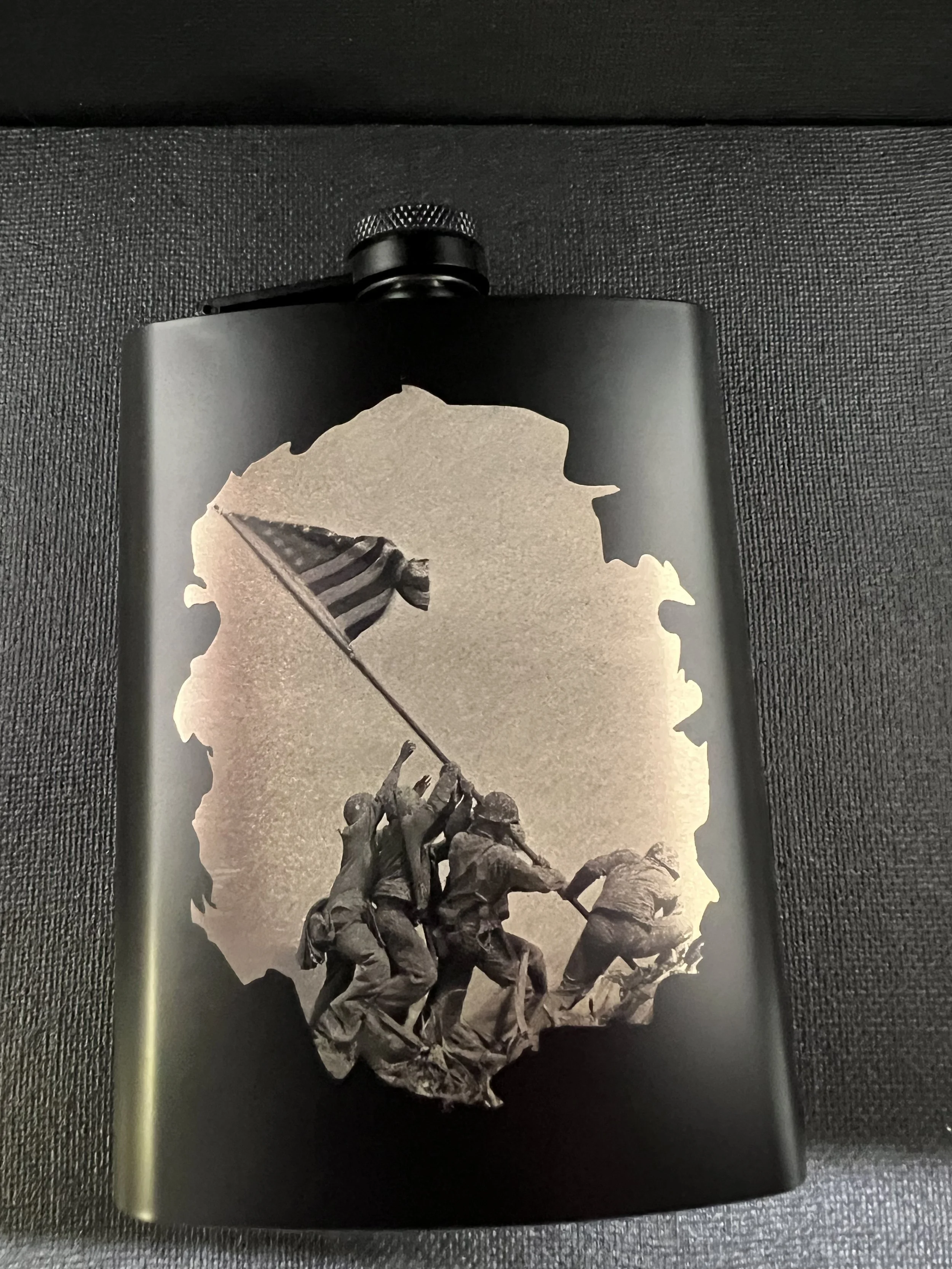 Black stainless steel hip flask engraved with the iconic Iwo Jima flag raising scene, shown against a textured dark background.