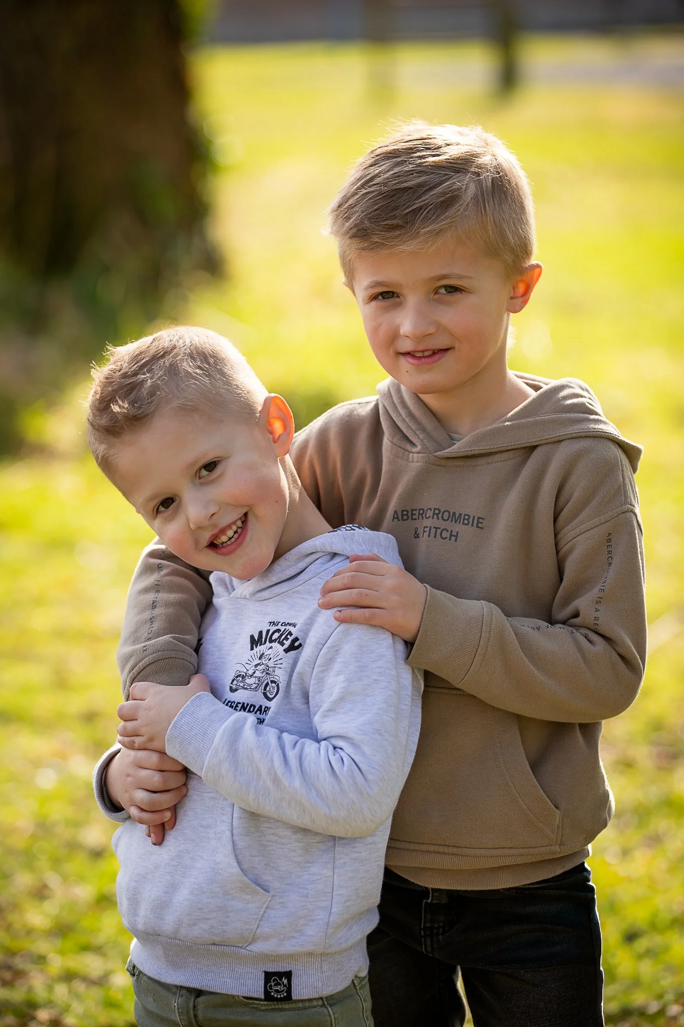 Deux jeunes garçons souriants en plein air lors d'une journée ensoleillée, l'un portant un sweat blanc et l'autre un hoodie beige, s'étreignant et posant pour la photo.