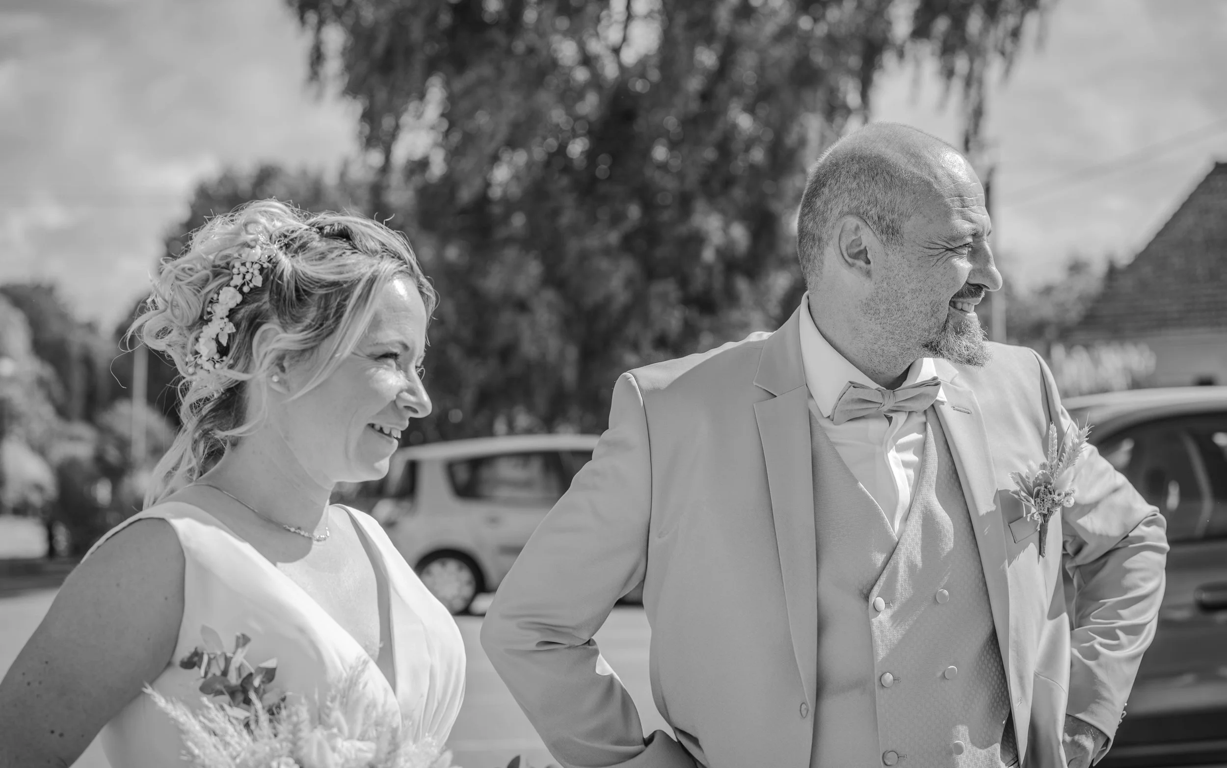 Deux personnes en costume de mariage, une femme et un homme, souriant dehors avec des voitures en arrière-plan.