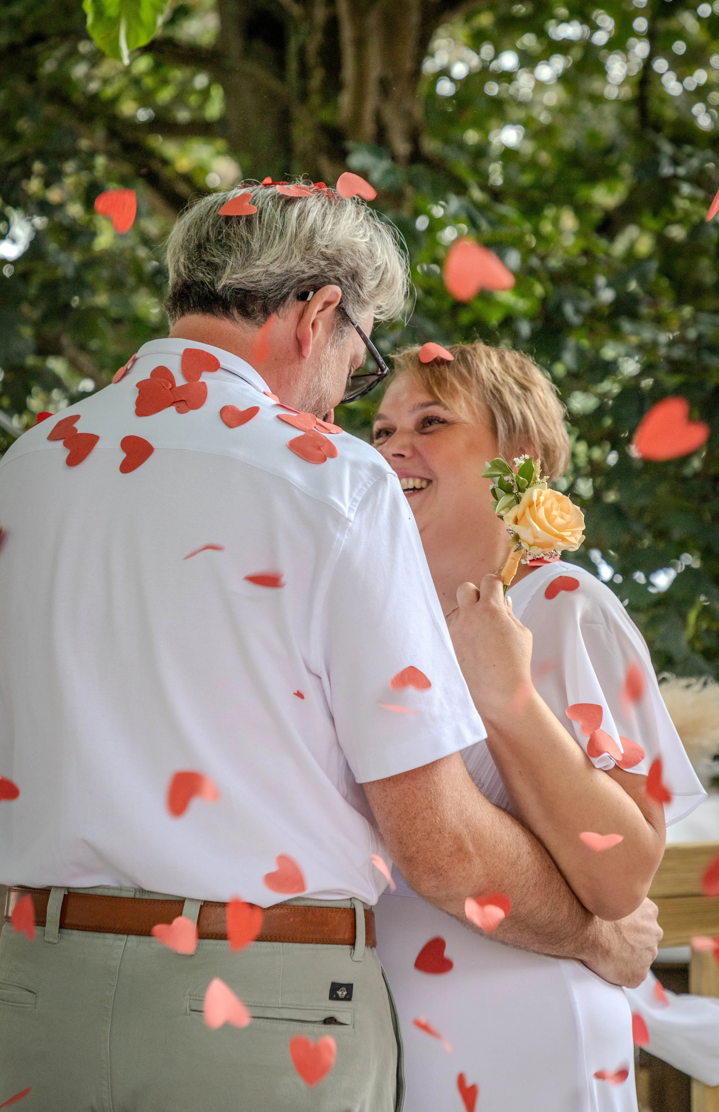 Un couple heureux lors d'un mariage, enveloppé de confettis en forme de cœurs, en train de s'aimer devant un arbre.