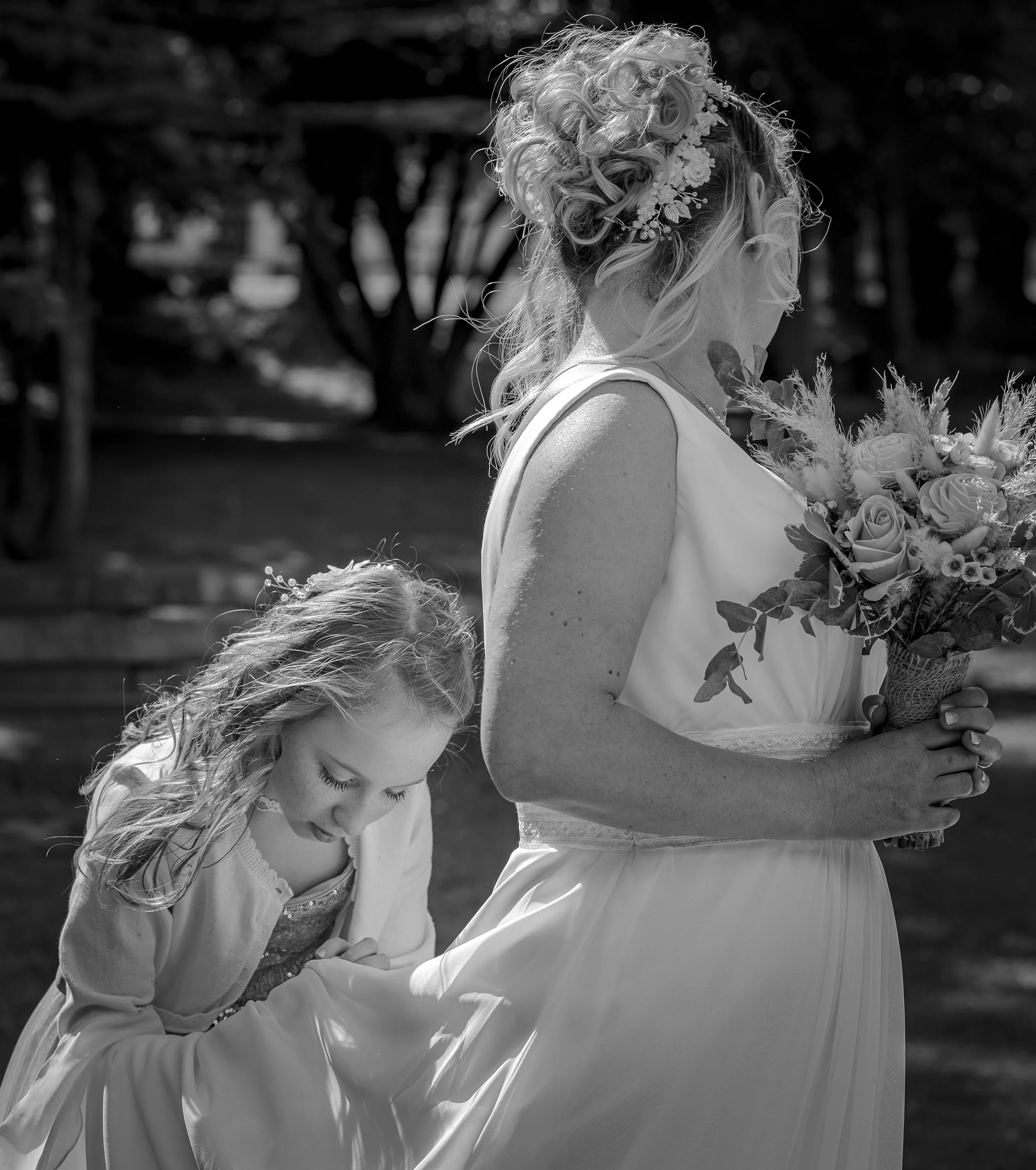 Une femme portant une robe de mariage tient un bouquet de fleurs, une jeune fille ajuste le bas de la robe dans un cadre extérieur