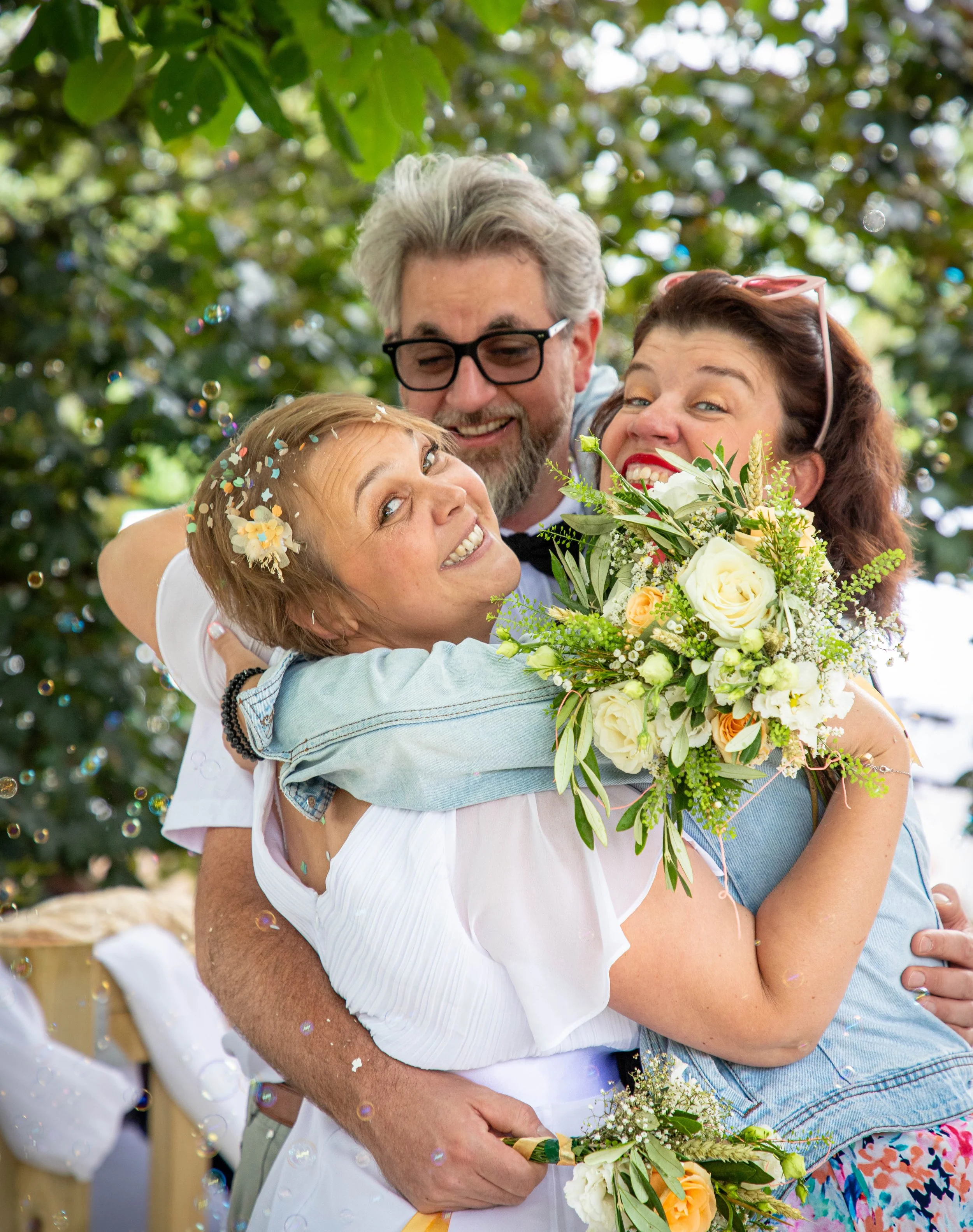Trois personnes souriantes en train de faire un câlin lors d'une célébration, avec des fleurs et des confettis. Arrière-plan de feuillage vert.