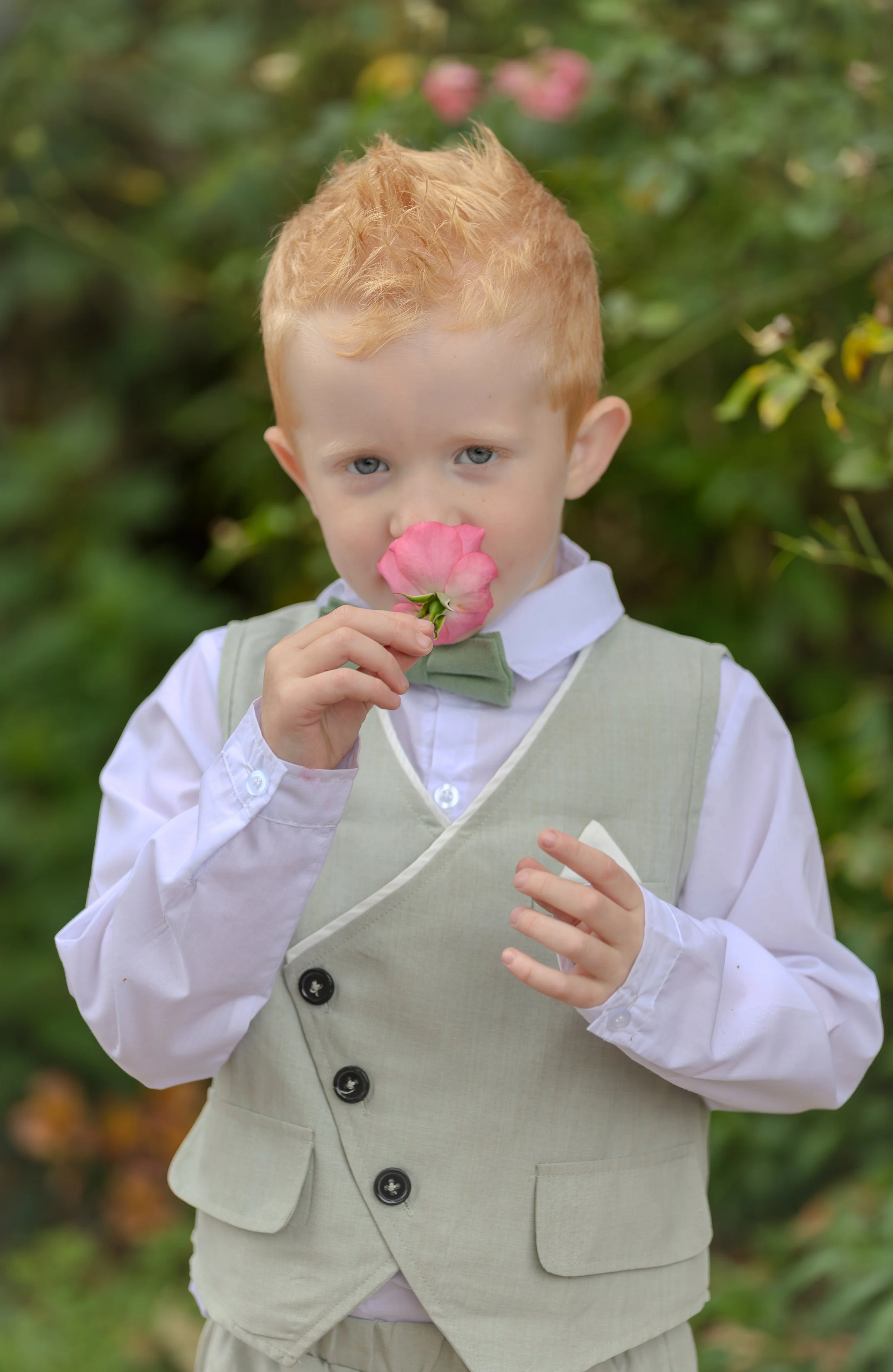 Un jeune garçon aux cheveux roux tient une fleur rose près de son visage, dans un jardin avec un fond de feuillage vert.
