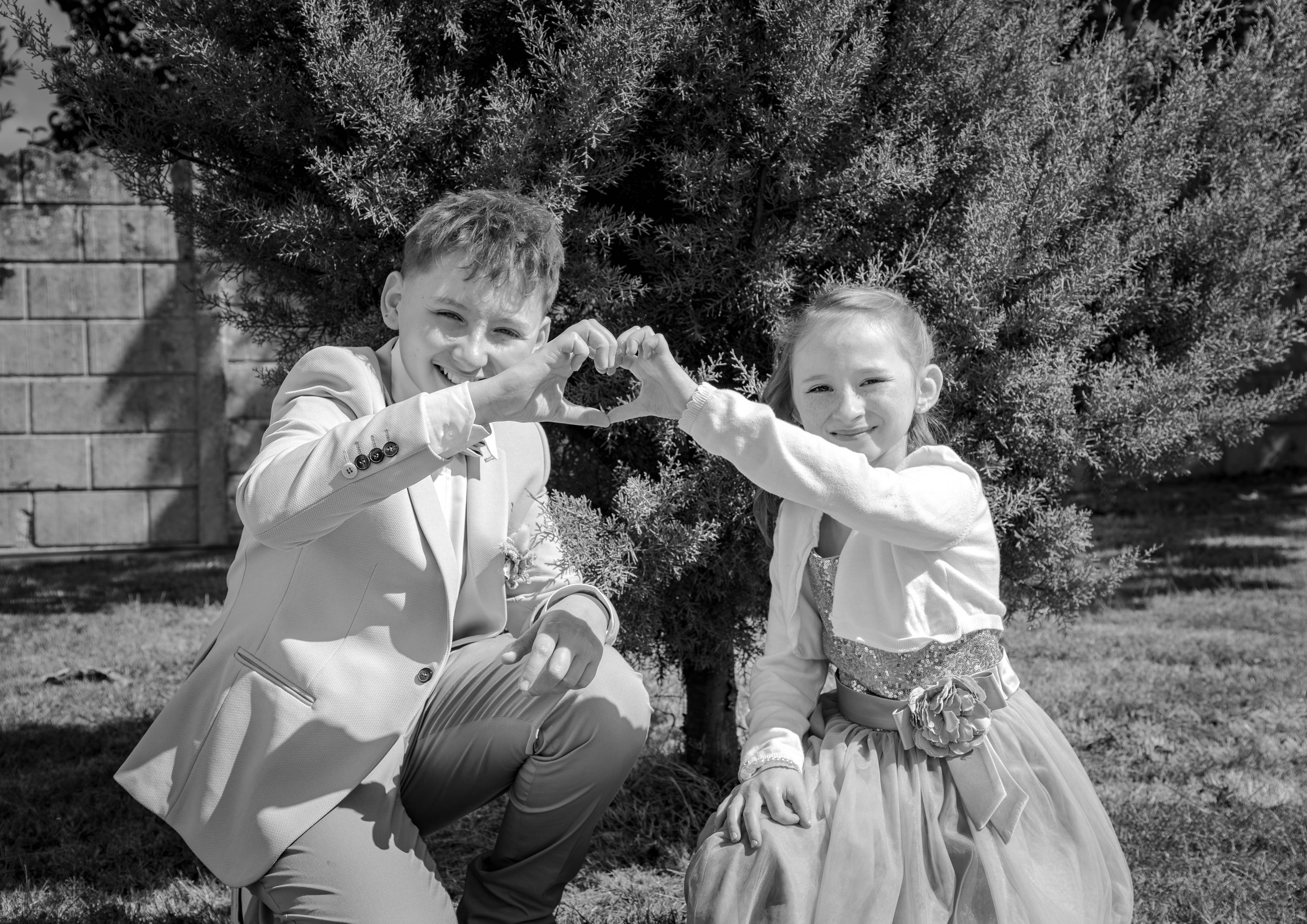 Deux enfants, un garçon et une fille, forment un cœur avec leurs mains devant un buisson d'arbuste, en plein air, en noir et blanc.