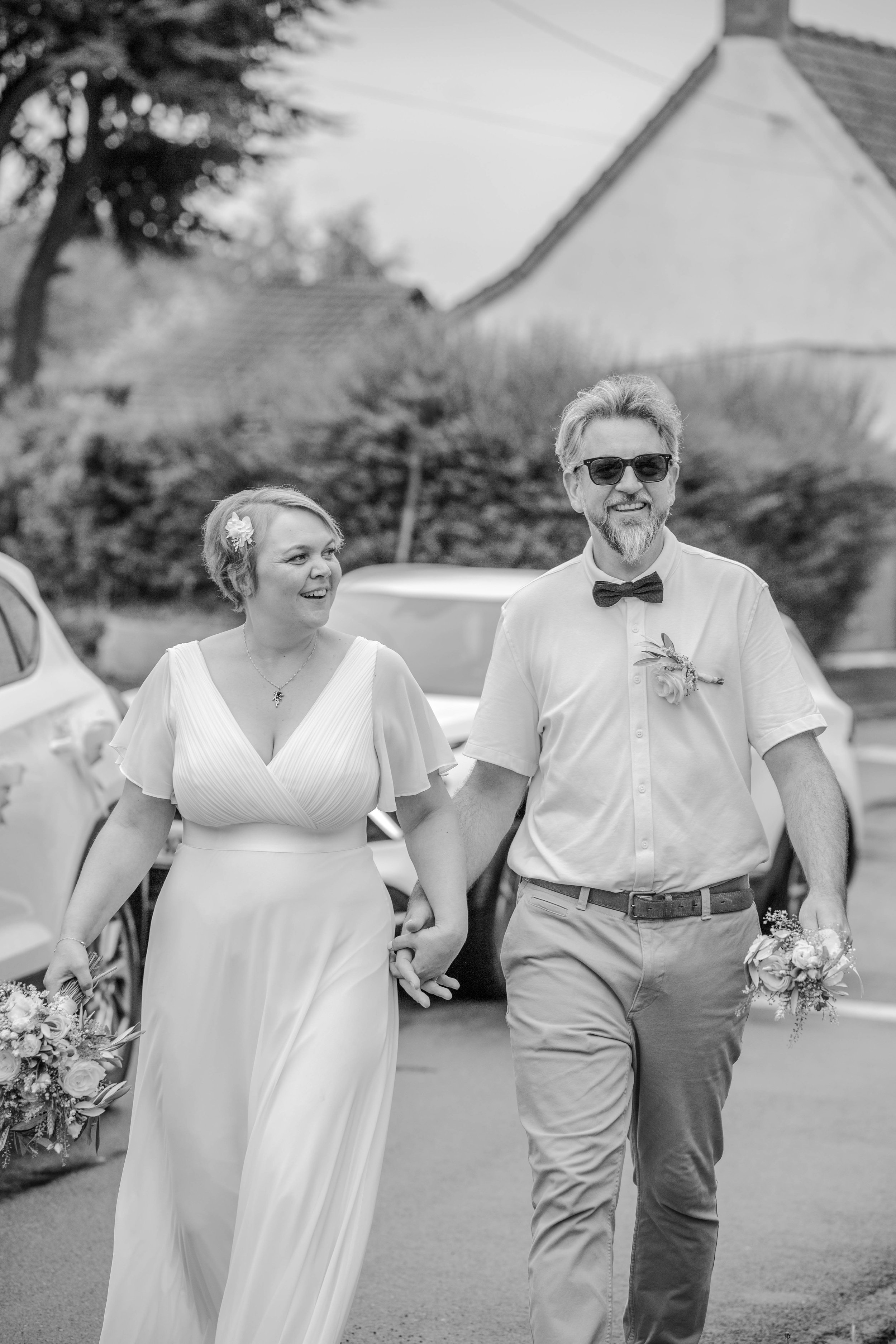 Un couple marchant main dans la main lors d'une cérémonie de mariage en extérieur, la femme porte une robe blanche et un bouquet, l'homme porte une chemise avec un nœud papillon, tous deux souriants.
