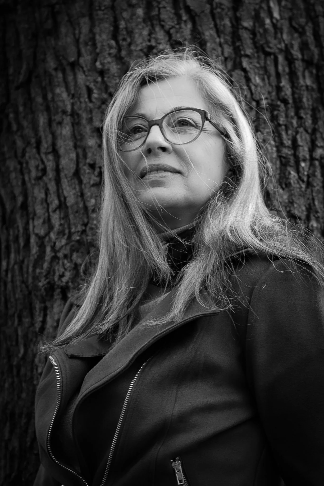Portrait noir et blanc d'une femme portant des lunettes, avec des cheveux longs et une veste en cuir, devant un arbre.