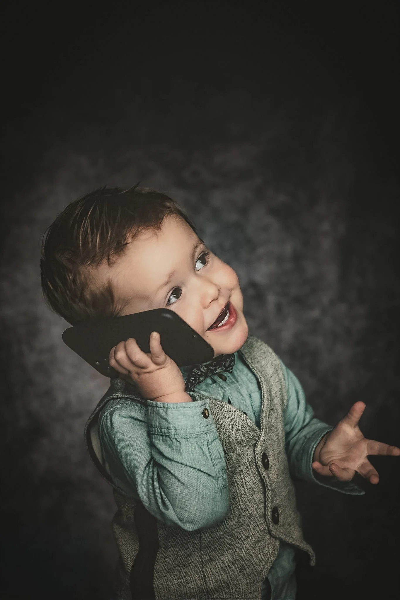 Un jeune garçon souriant, parlant au téléphone et faisant un signe de paix avec la main, contre un fond sombre.