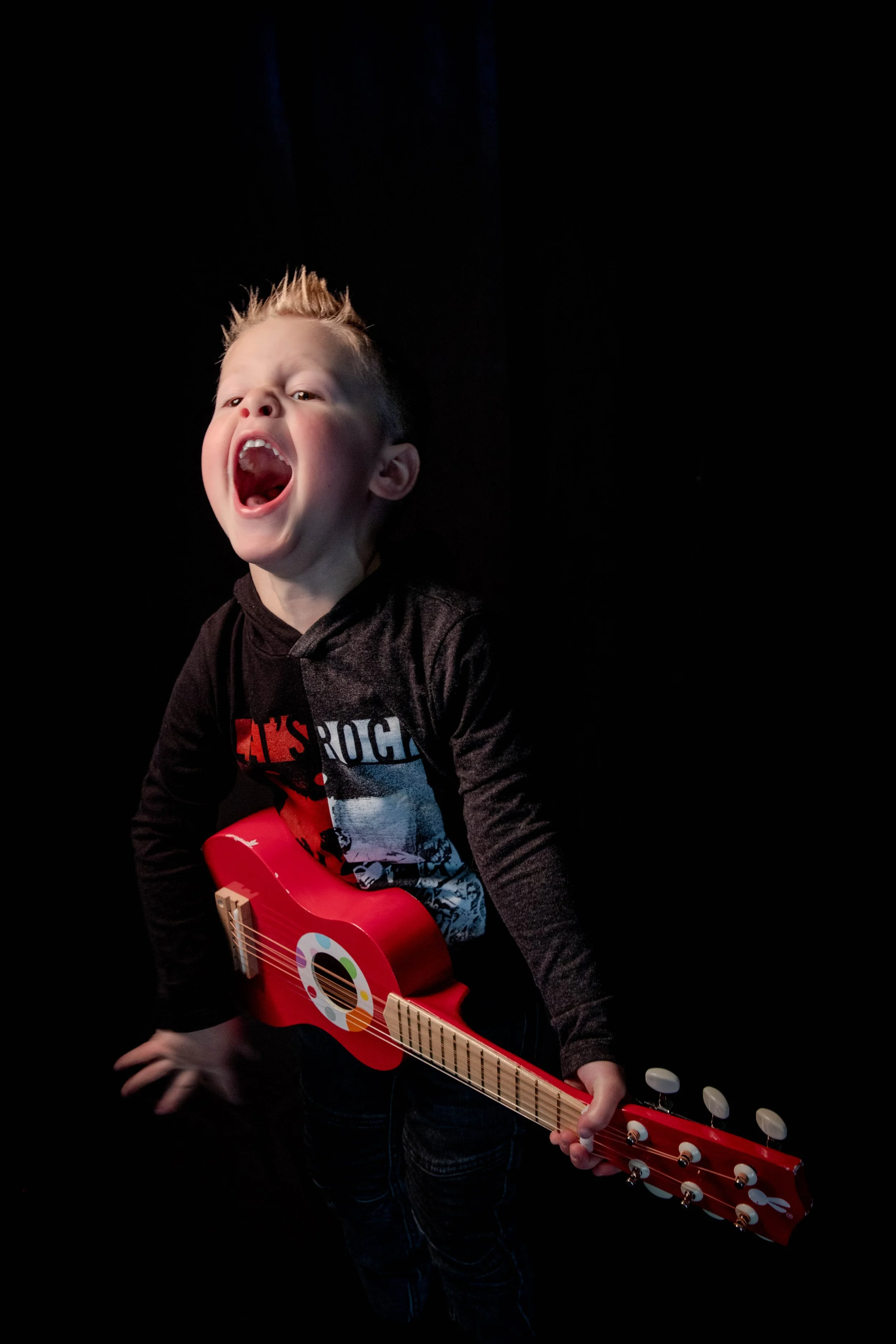 Un jeune garçon avec des cheveux en épis, chantant ou criant avec une guitare rouge, portant un t-shirt noir avec un motif.