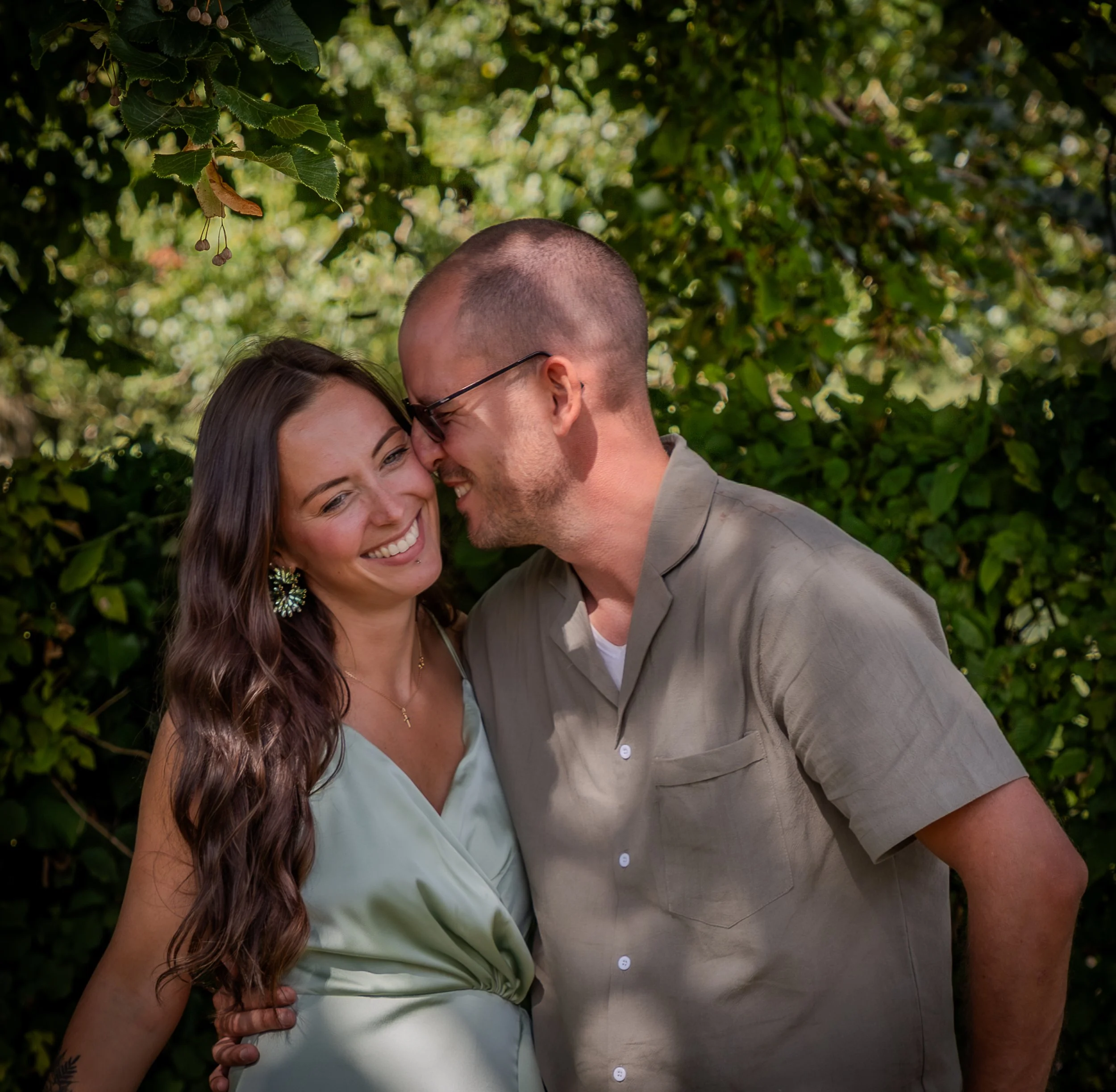 Un couple souriant et riant ensemble, en contact proche, dans un environnement extérieur verdoyant.