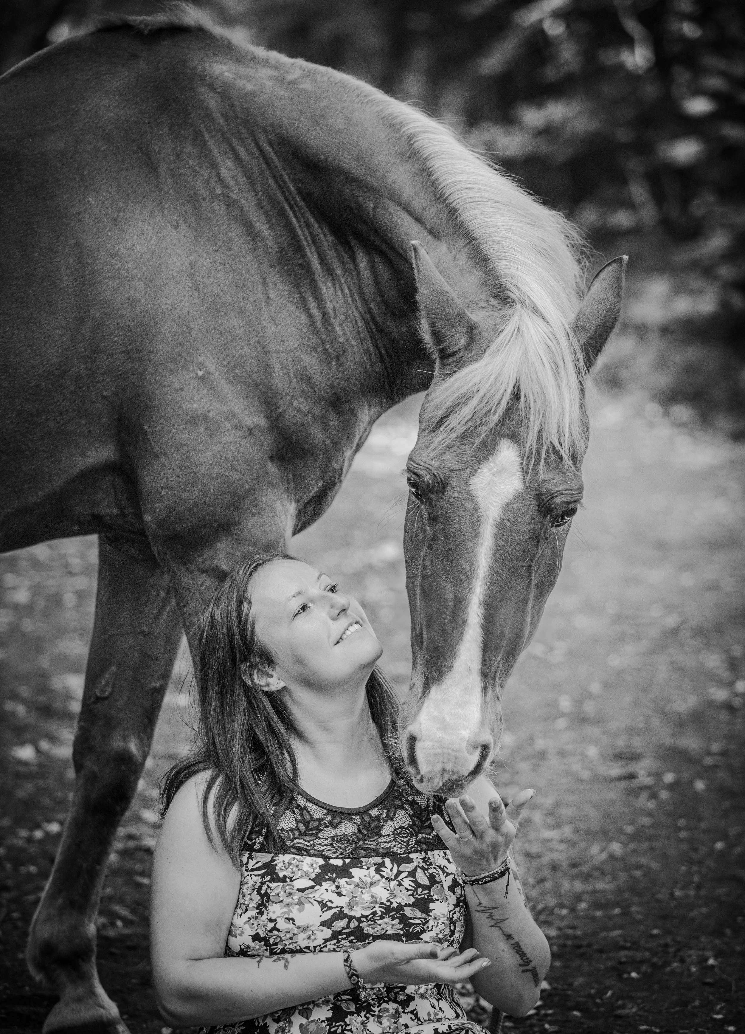 Une femme souriante regarde un cheval, qui penche la tête vers elle, dans un cadre extérieur naturel. La photo est en noir et blanc.