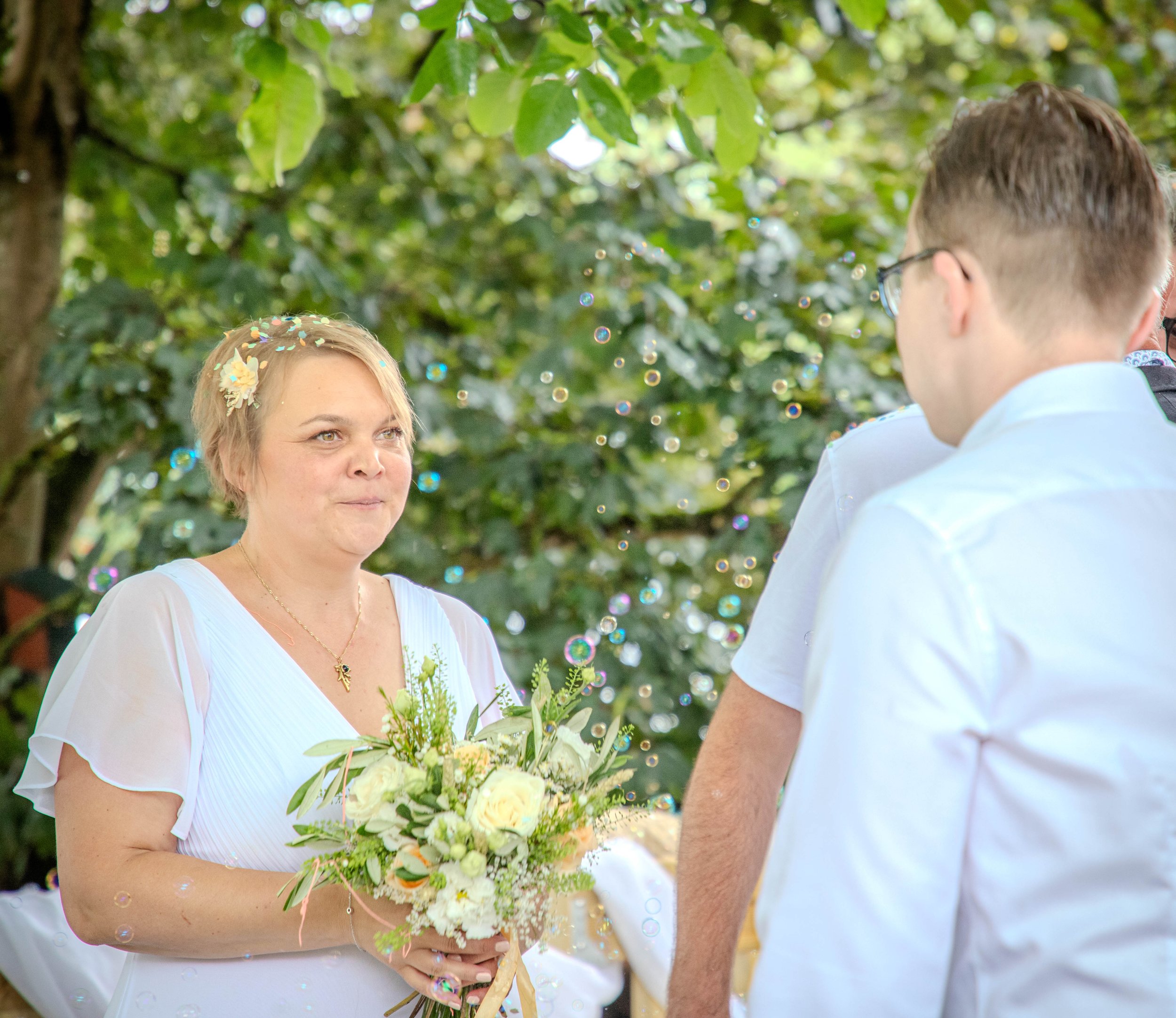 Une femme tenant un bouquet de fleurs lors d'un mariage, discutant avec un homme portant des lunettes. Contexte extérieur avec des feuilles vertes.