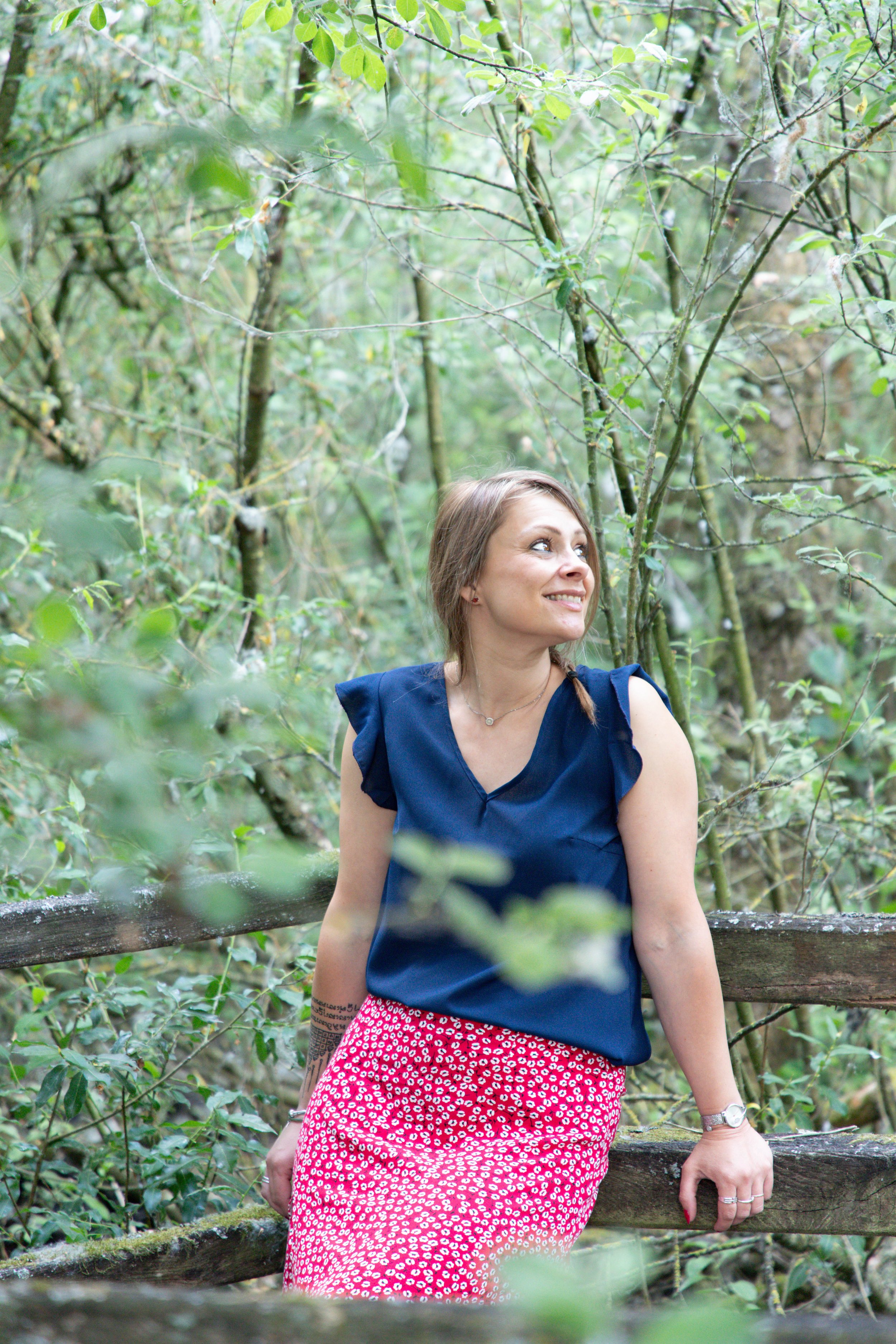 Femme souriante avec un haut bleu et une jupe rouge à pois, en train de se détendre dans une forêt, appuyée sur une barrière en bois.