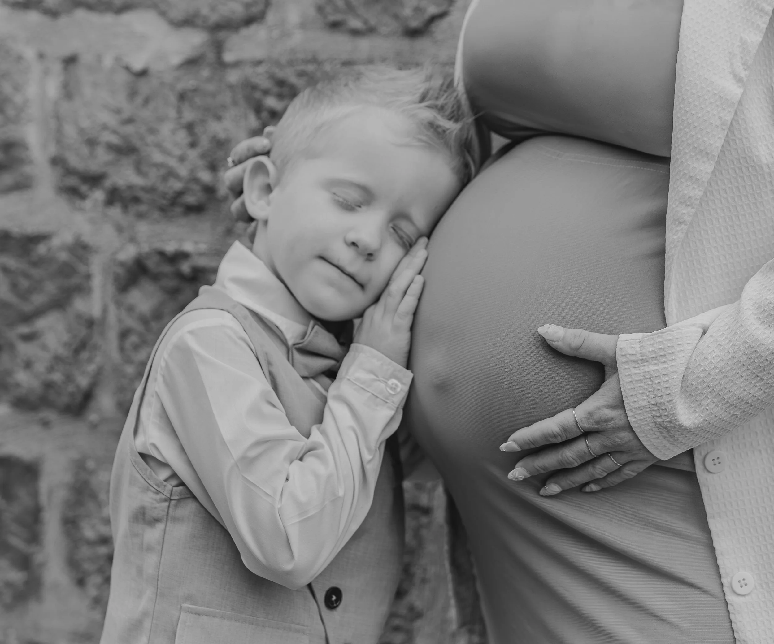 Un jeune enfant fait un câlin à un ventre rond de femme enceinte, avec un sourire doux, en noir et blanc.