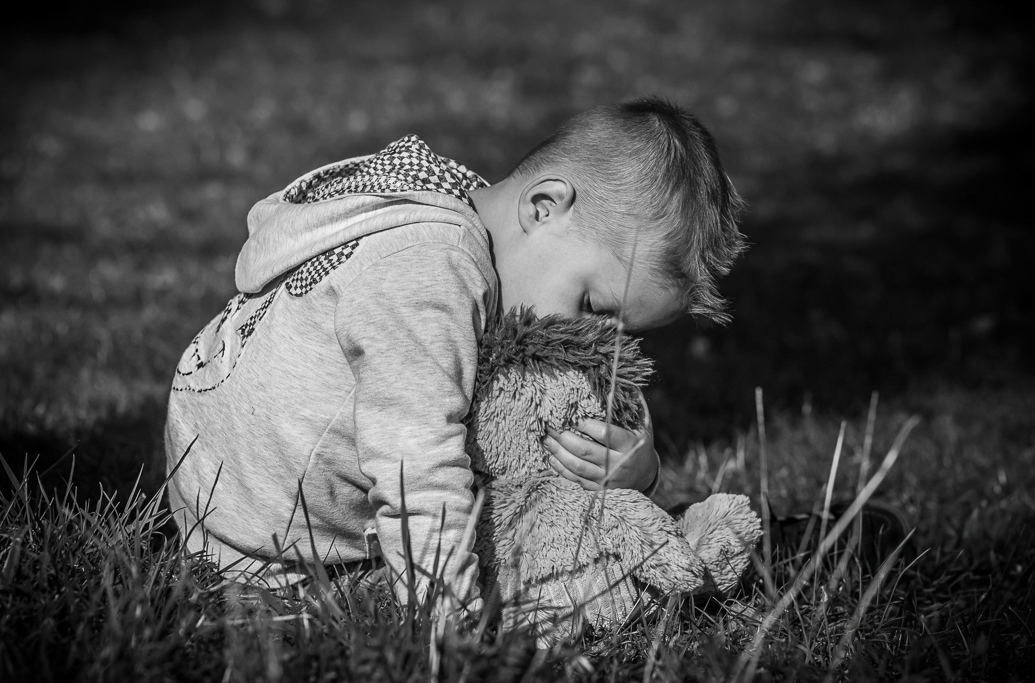 Un jeune garçon embrasse tendrement une peluche en plein air, en noir et blanc.