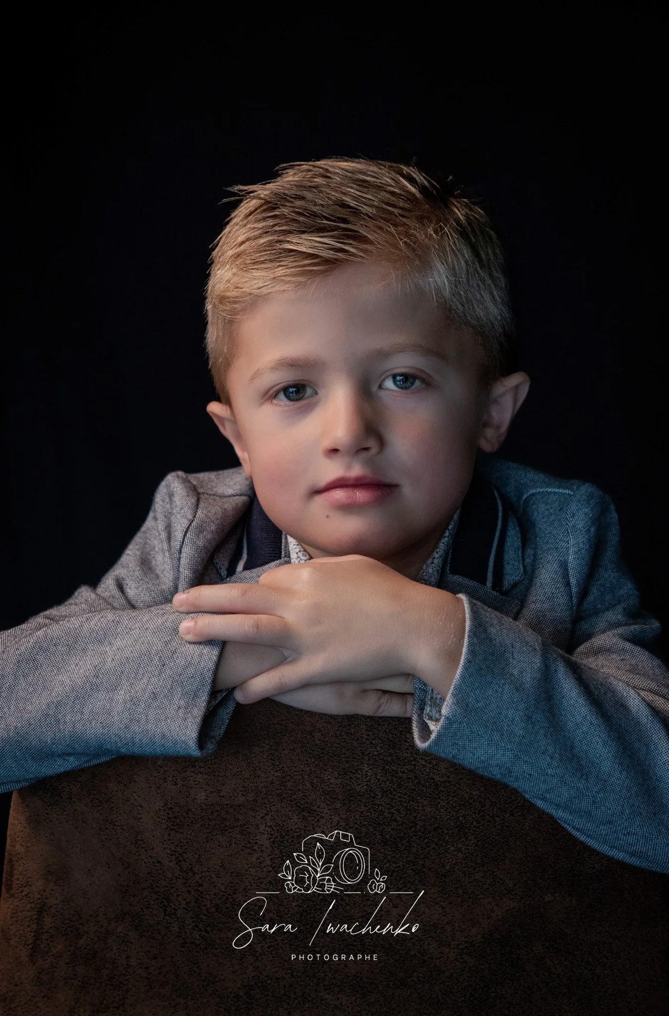 Jeune garçon blond aux yeux bleus portant un costume gris, posé sur une surface sombre, regardant directement l'objectif.