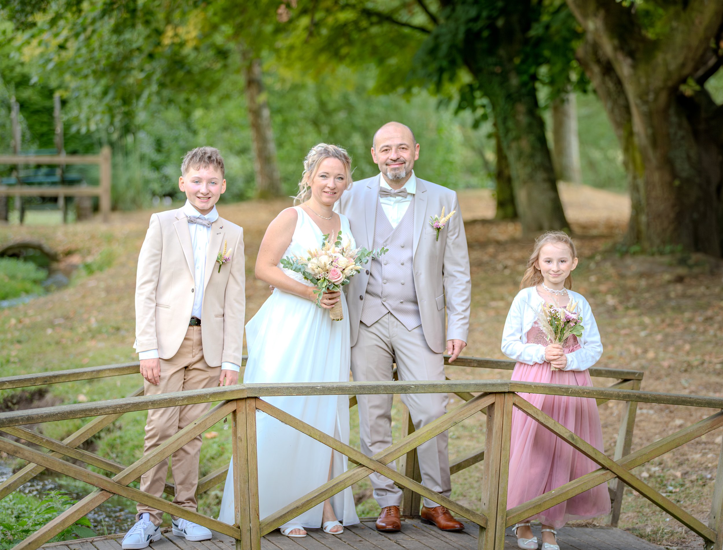Un groupe de cinq personnes, un couple avec deux enfants, pose pour une photo lors d'un mariage en plein air devant un décor naturel avec des arbres et une petite rivière en arrière-plan.