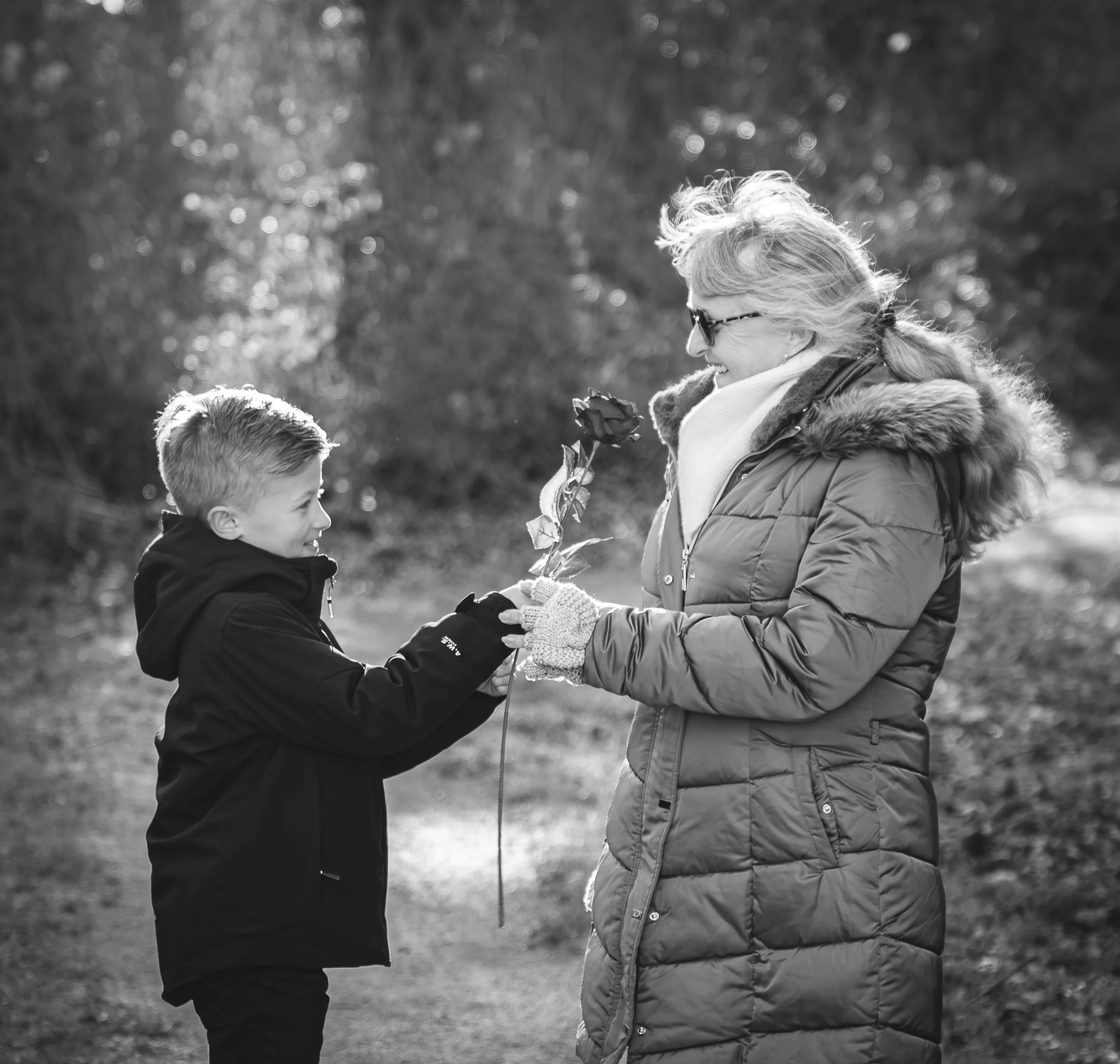Une femme donne une rose à un garçon dans un parc, tous deux souriants, en pleine nature, en noir et blanc.