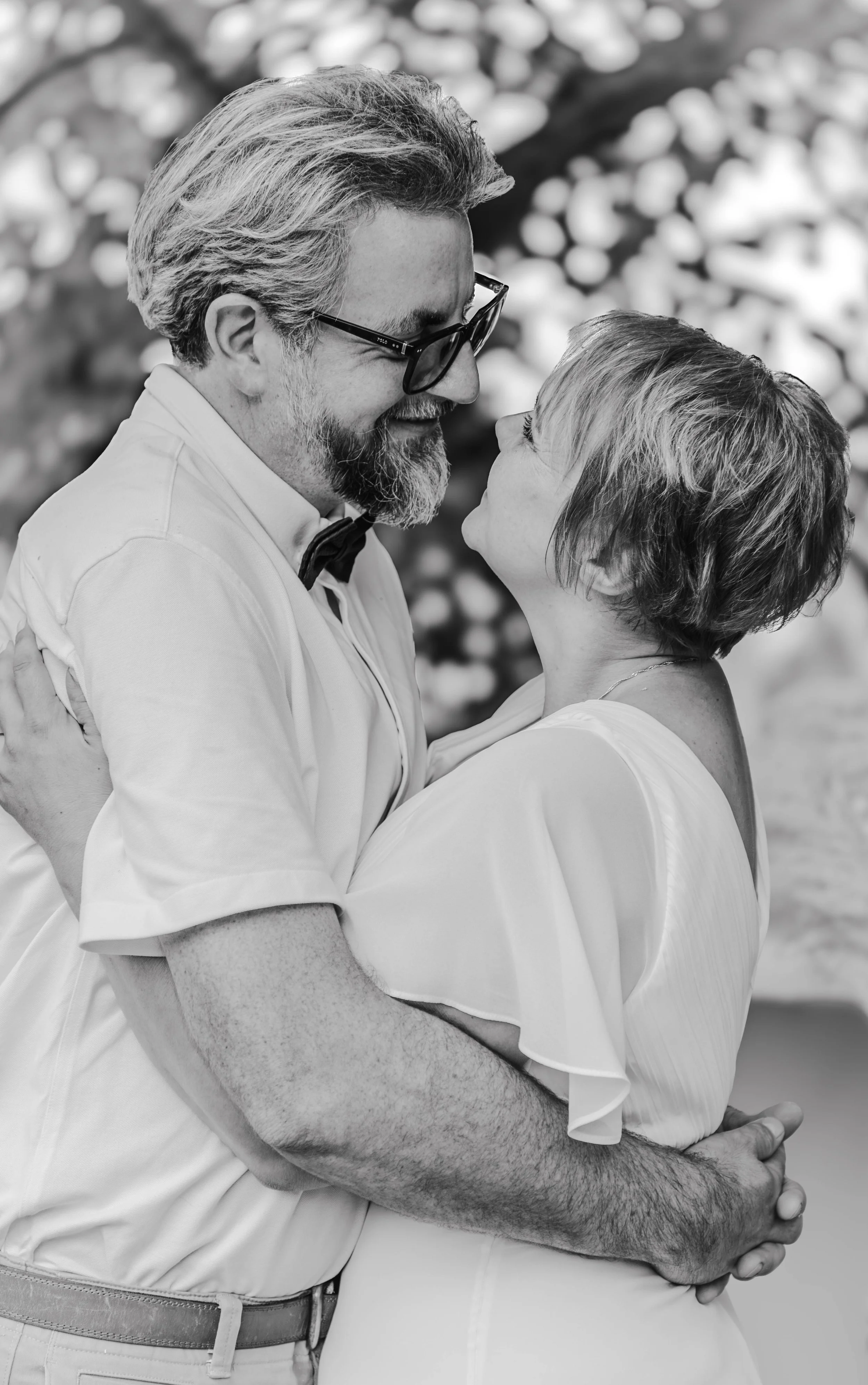 Un couple en train de danser, regardant attentivement l'un l'autre, avec un arrière-plan flou d'arbres et feuilles. La photo est en noir et blanc.