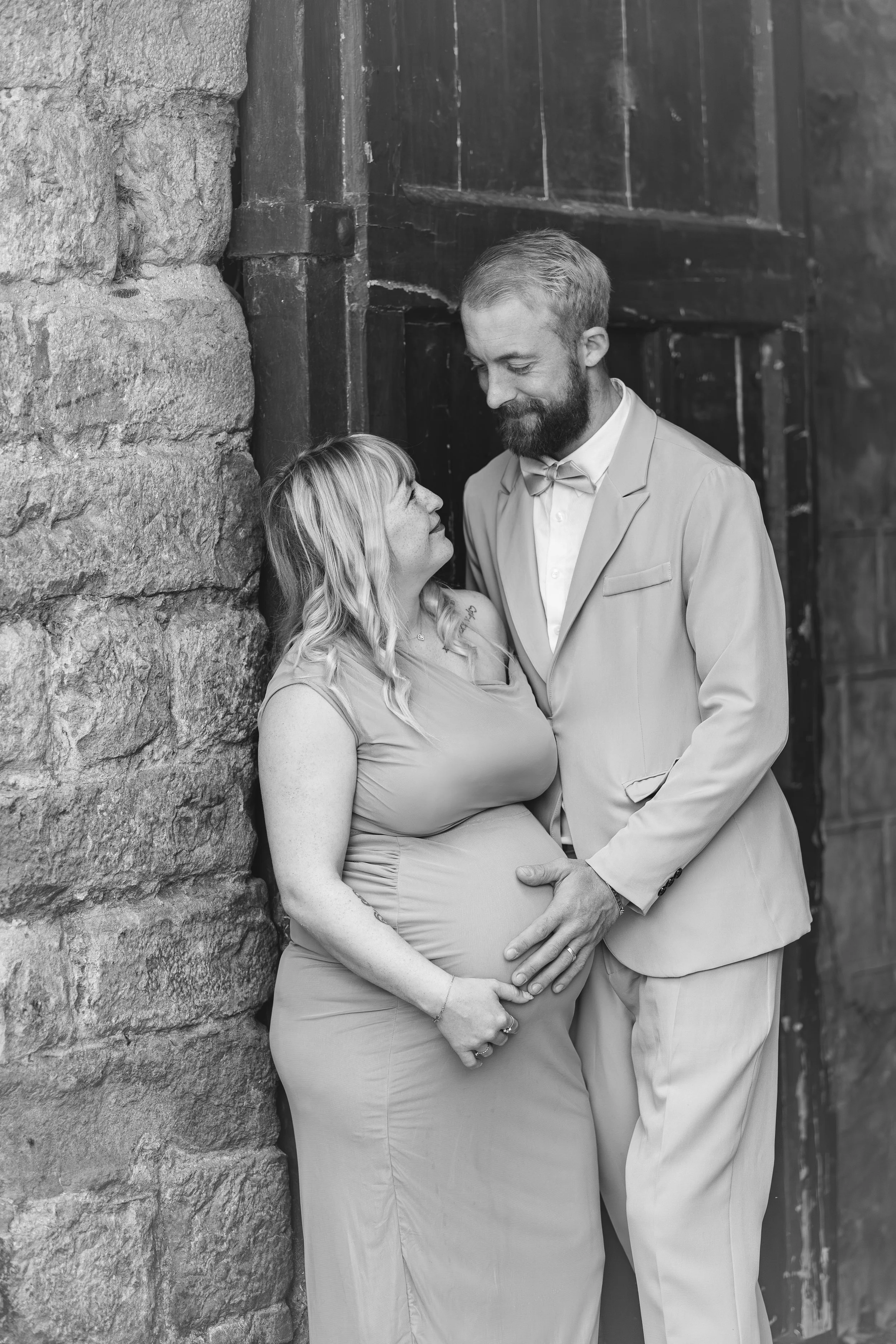 Une femme enceinte et un homme sourient en se regardant, debout contre un mur en pierre, en noir et blanc.
