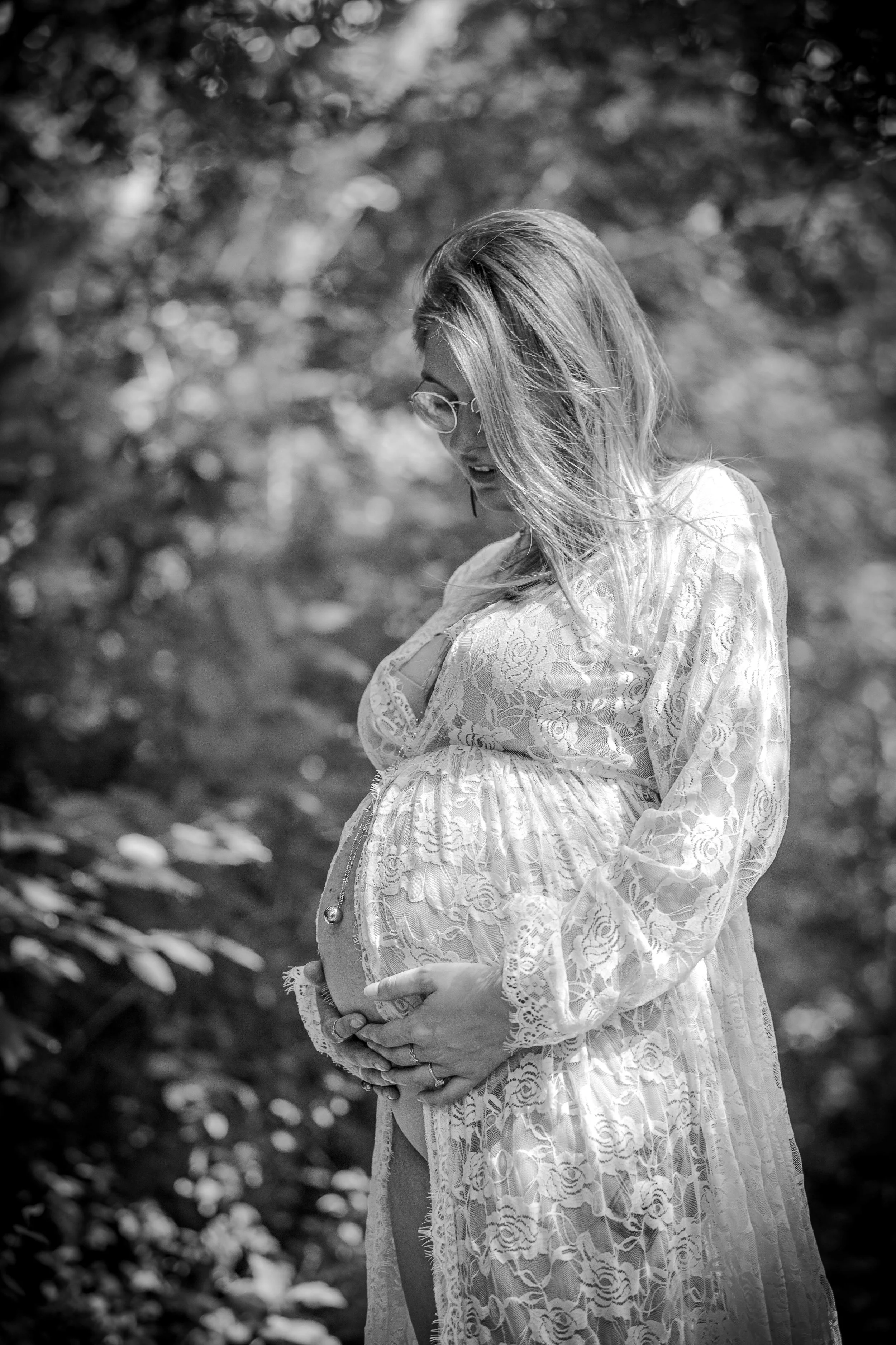 Une femme enceinte portant une robe en dentelle, tenant son ventre et portant des lunettes de soleil, dans un environnement extérieur avec des arbres en arrière-plan, en noir et blanc.