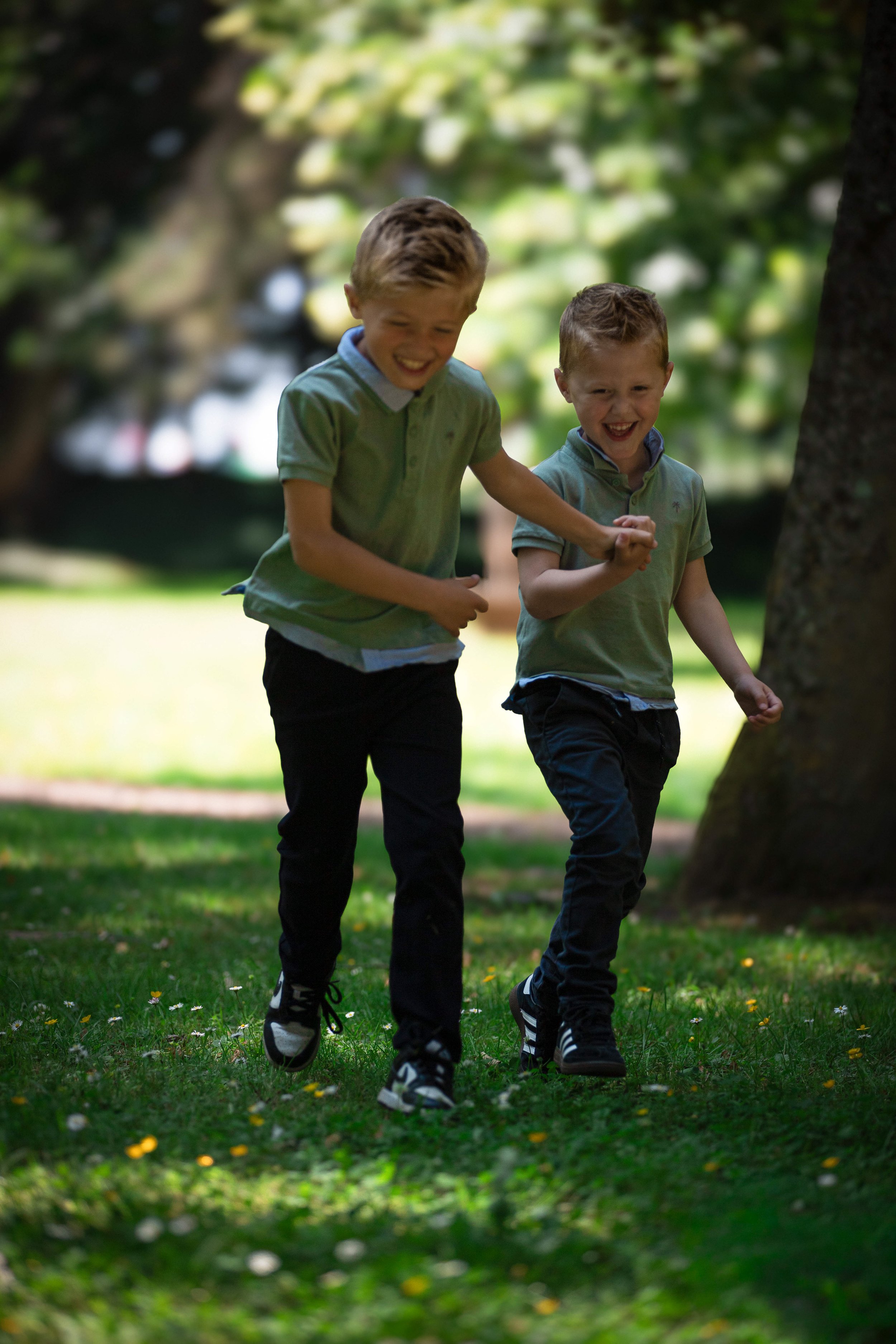 Deux jeunes garçons jouent et rient dans un parc ensoleillé, se tenant main dans la main, entourés d'arbres et d'herbe verte.