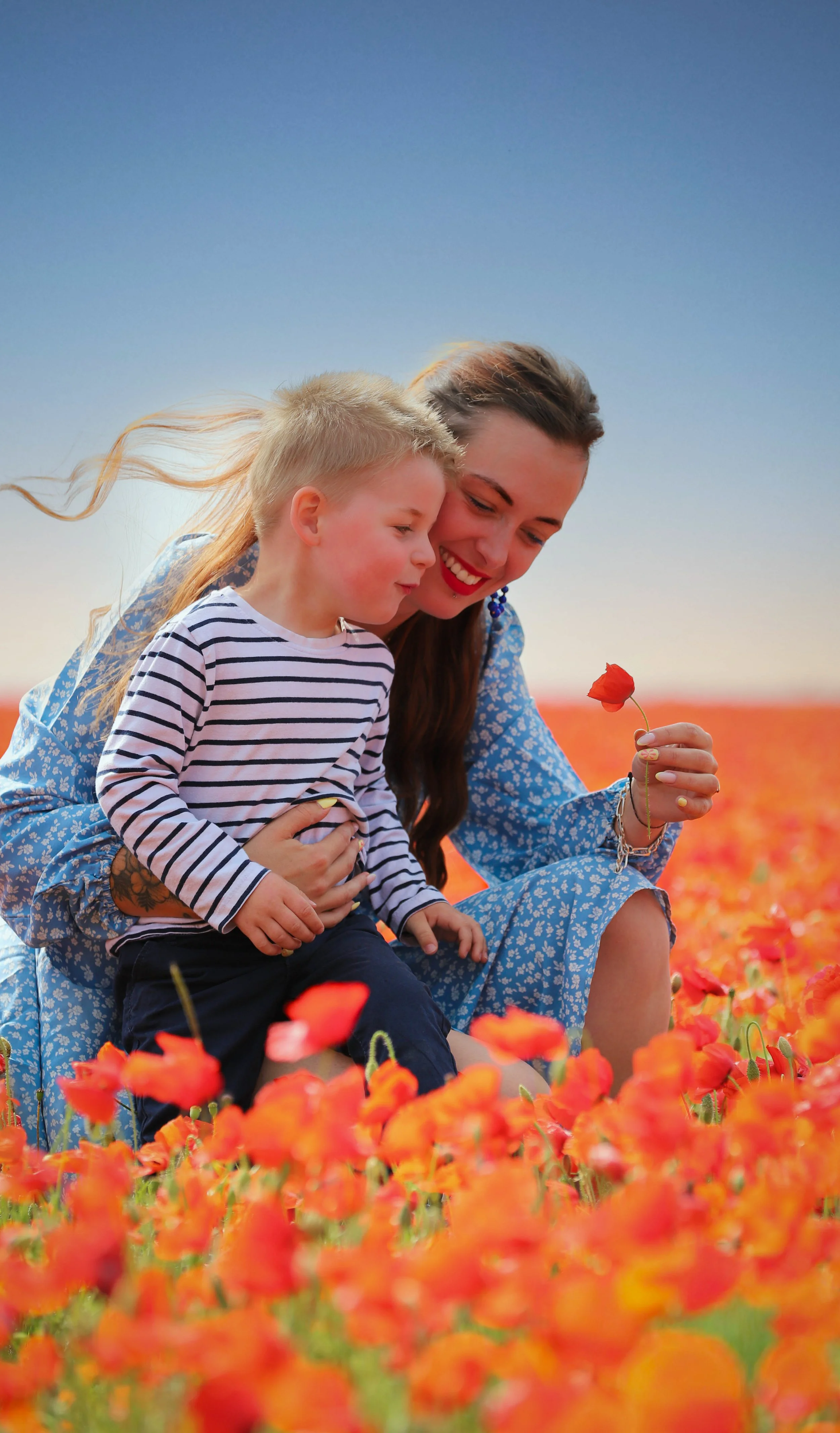 Une femme et un jeune garçon assis dans un champ de coquelicots, la femme tient une fleur et sourit, tous deux regardant la fleur dans la main de la femme.