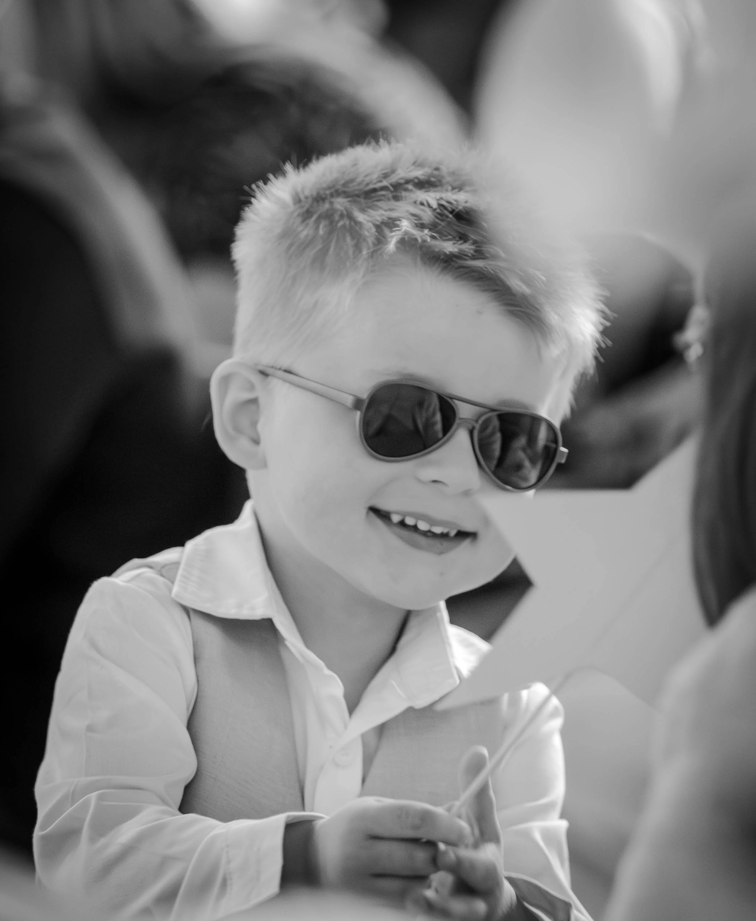 Un garçon souriant portant des lunettes de soleil, habillé d'une chemise blanche, qui regarde une feuille qu'il tient à la main.