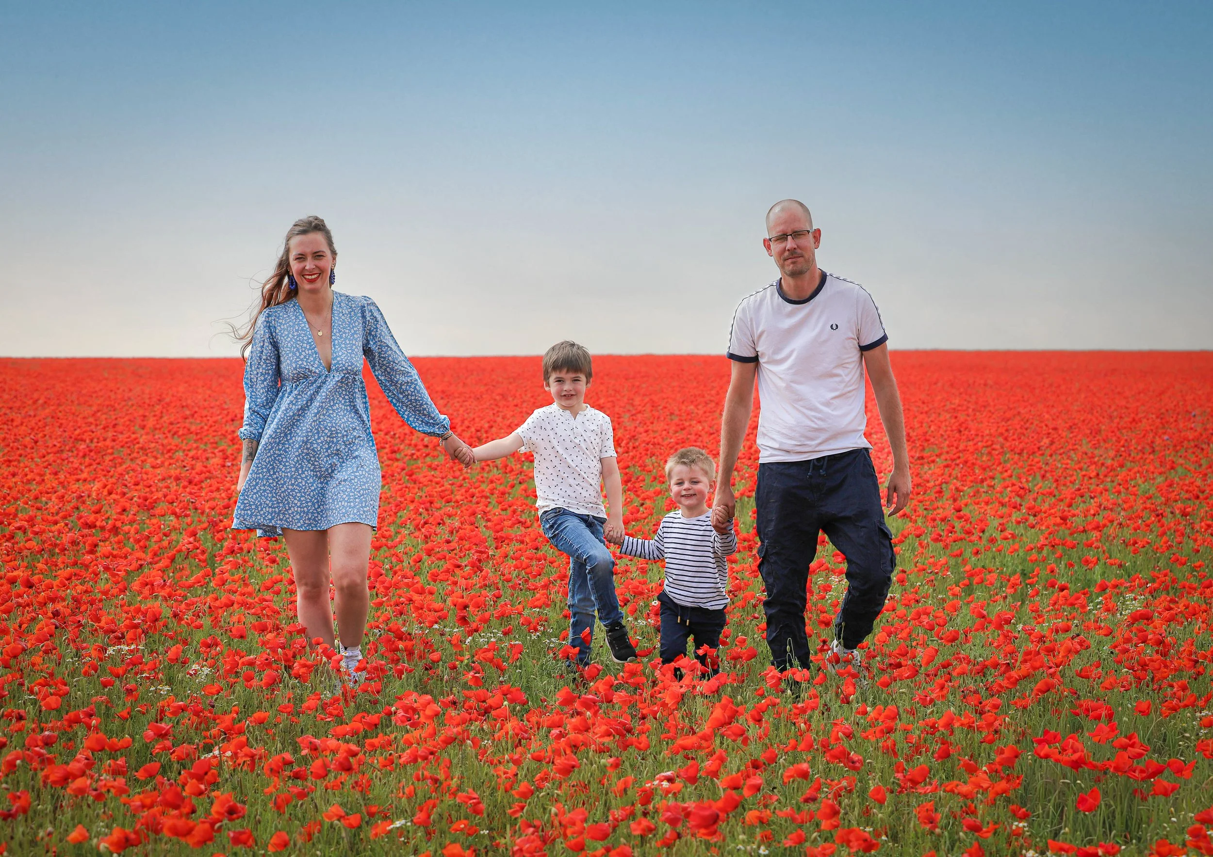 Une famille de quatre personnes, deux adultes et deux enfants, marche dans un champ de coquelicots rouges sous un ciel clair.