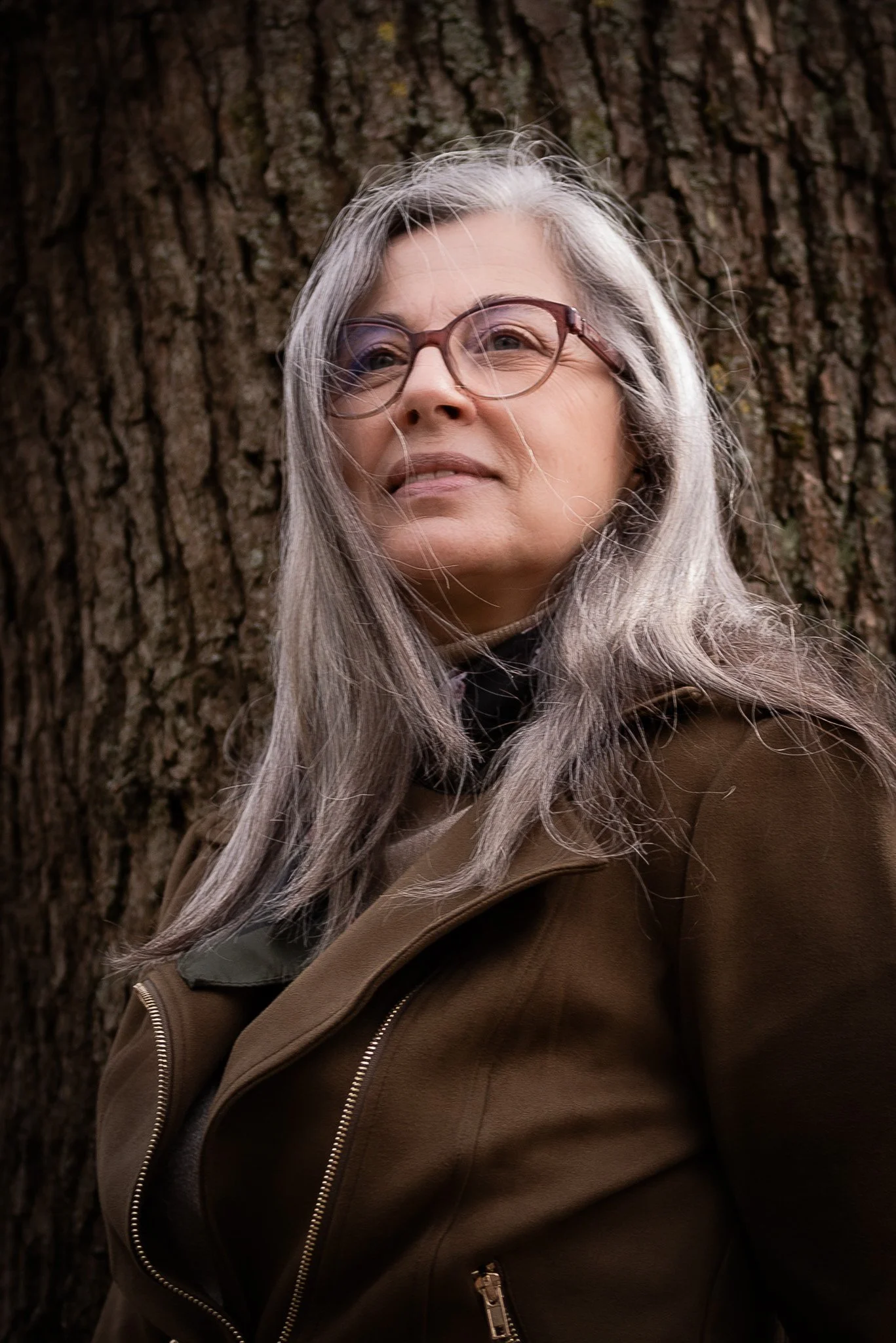 Une femme âgée, aux cheveux longs gris, portant des lunettes et un manteau marron, se tient devant un arbre.